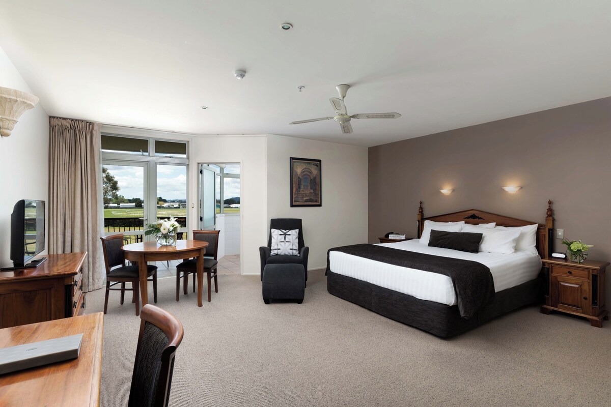 Quarto de hotel com cama, escrivaninha e cadeira, televisão, mesa de jantar com cadeiras e vista para o exterior.