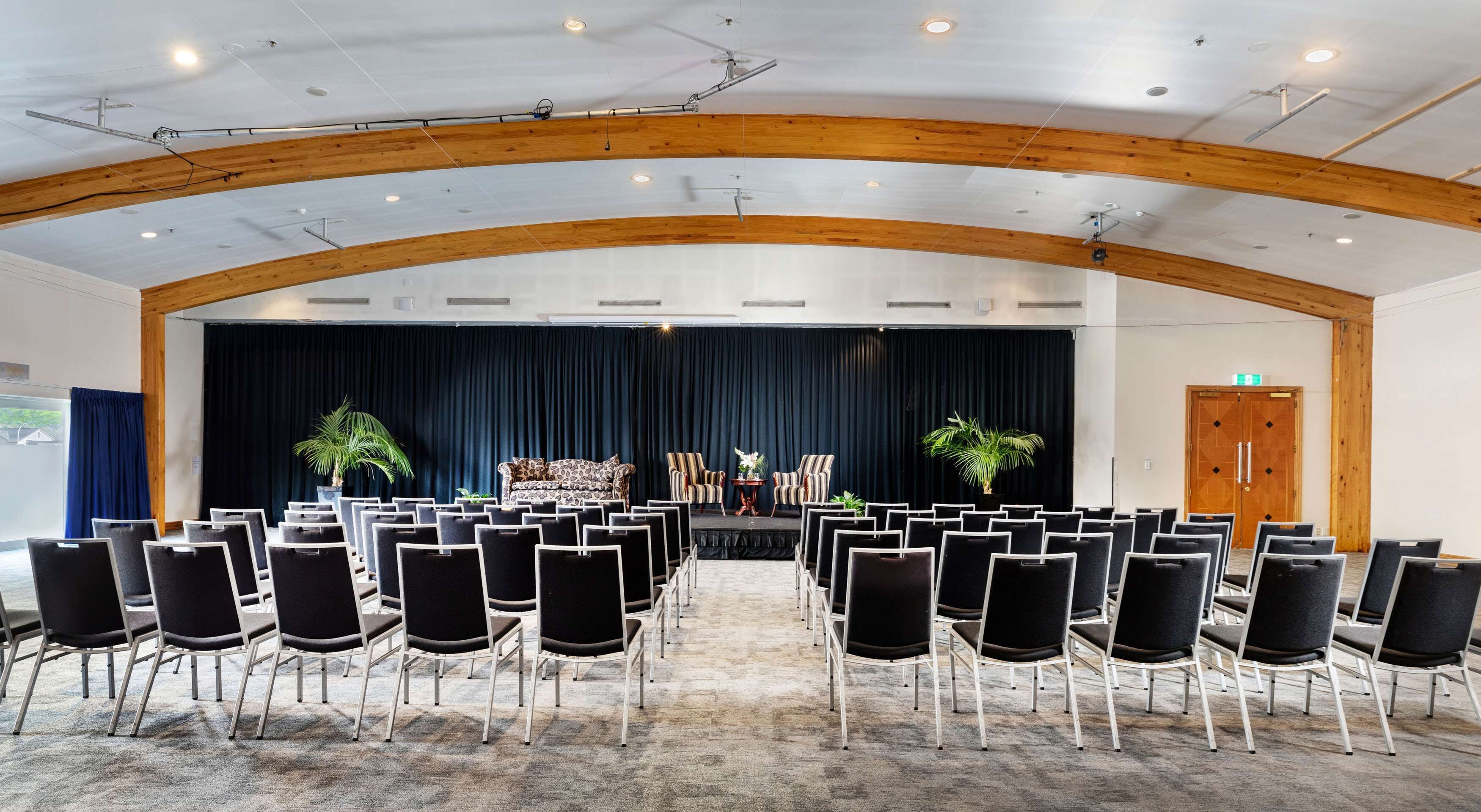 A conference room with rows of chairs, a stage with curtains, and wooden beams on the ceiling.