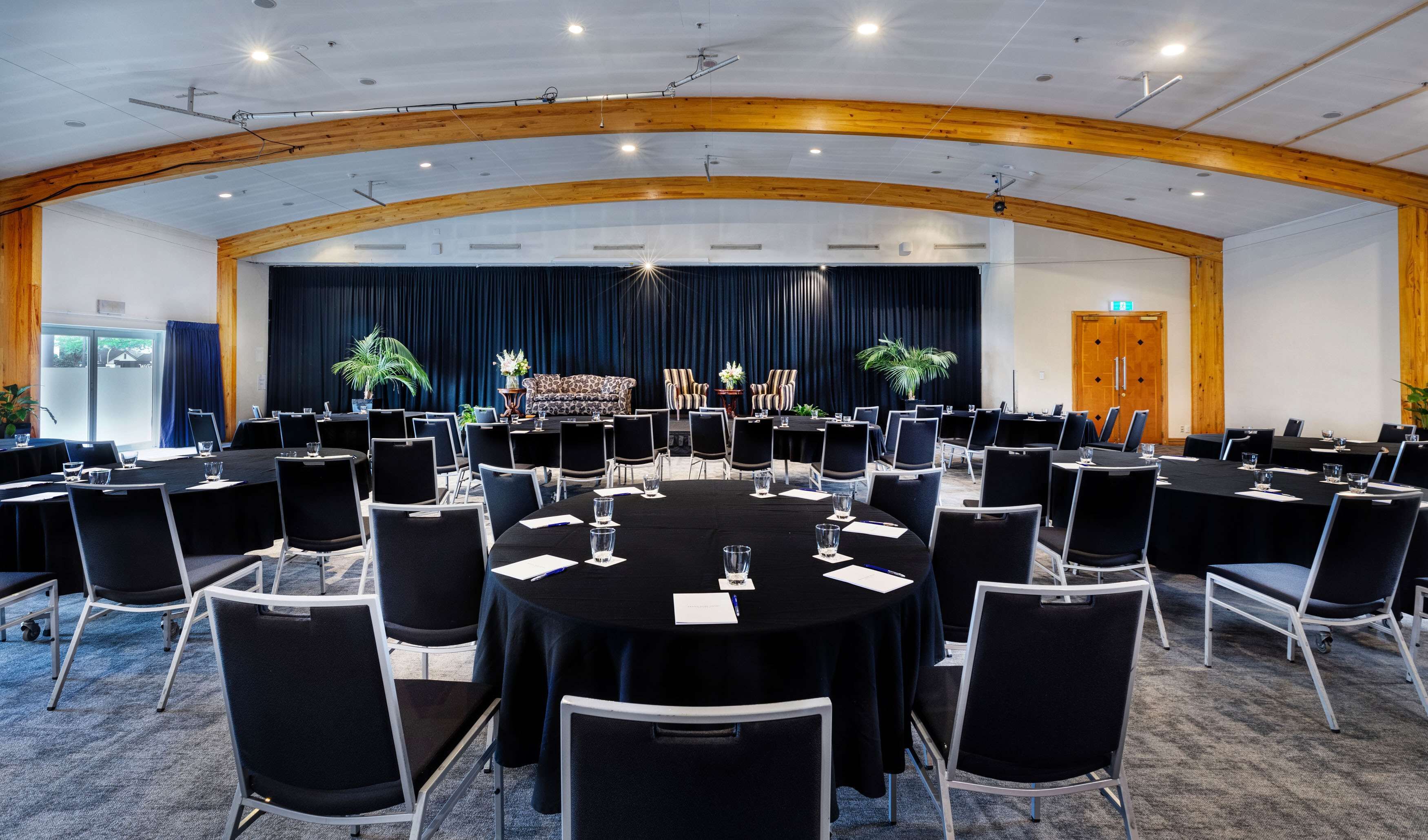 Sala de conferências com mesas redondas, cadeiras pretas e palco com cortinas.
