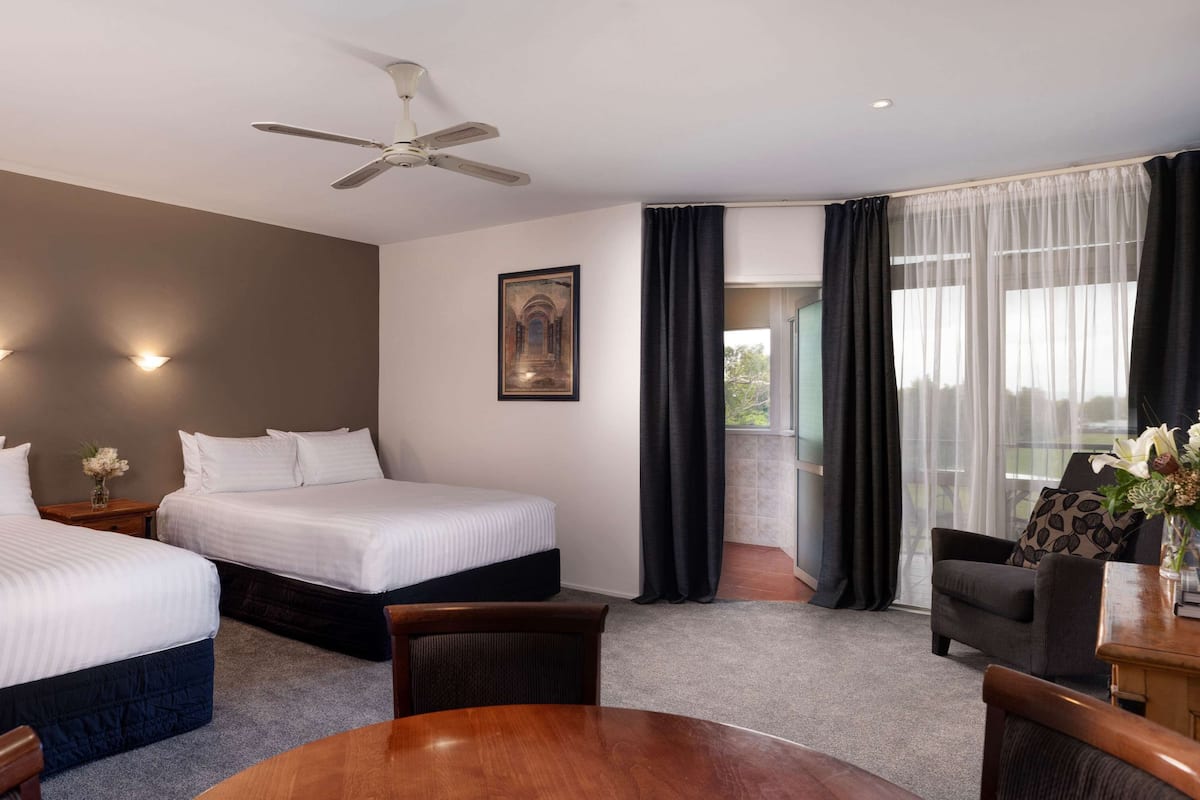 A hotel room with two beds, a ceiling fan, a painting, a view through a window, and a dining area with a table and chairs.