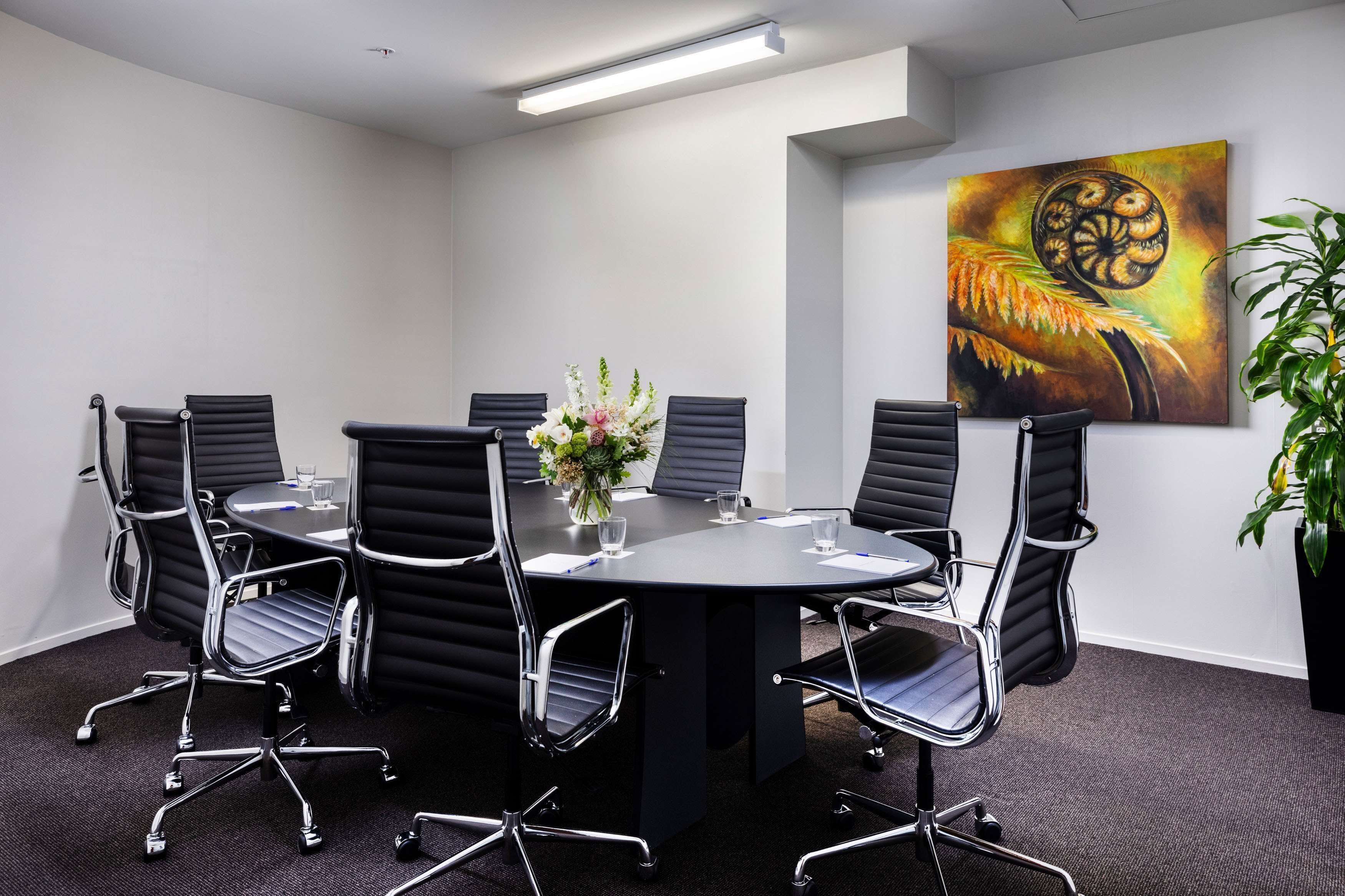 Une salle de conférence avec une table ronde, des fauteuils en cuir noir, un vase rempli de fleurs et un tableau accroché au mur.