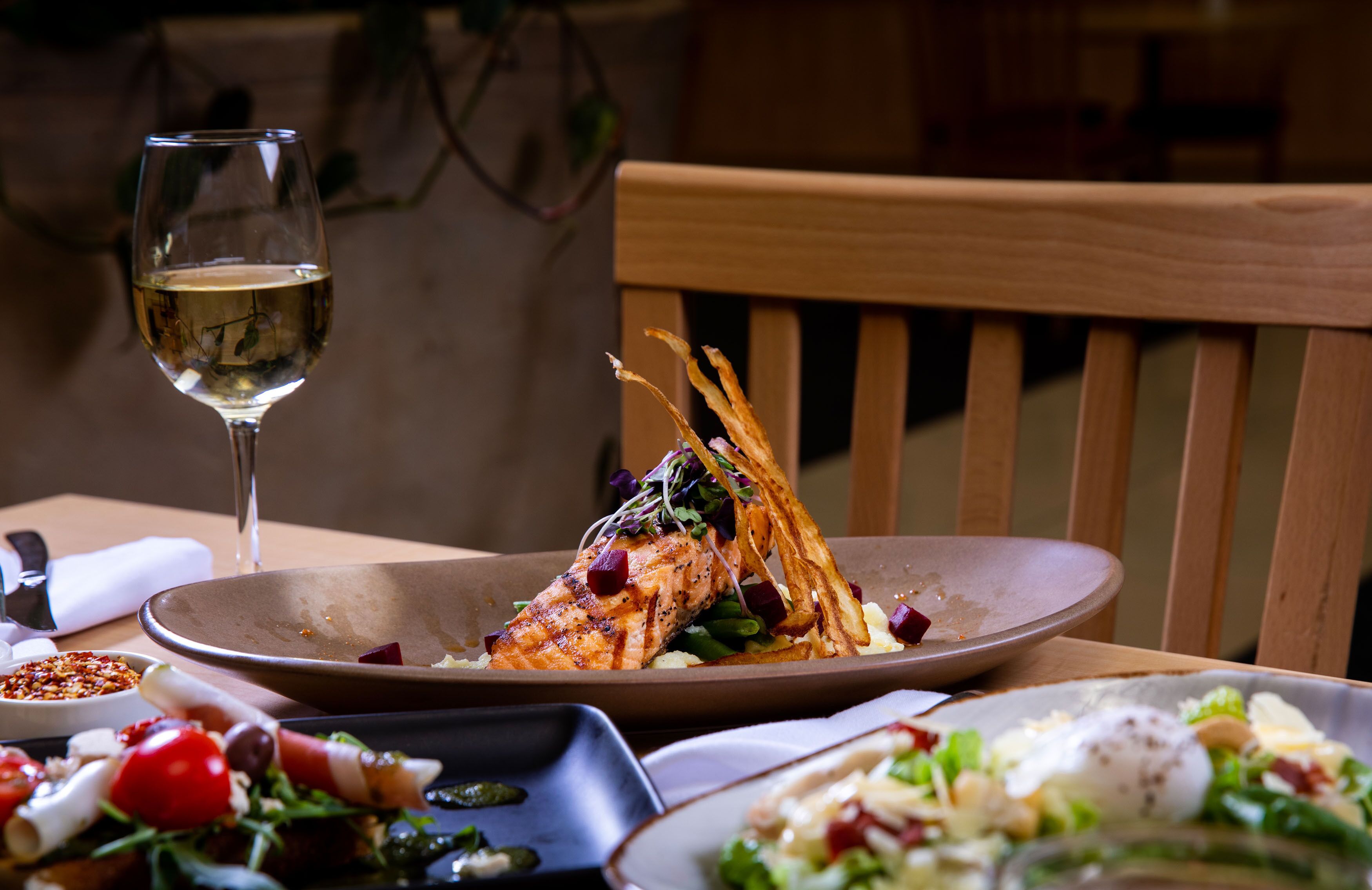 Una mesa de comedor con un plato de comida, una copa de vino blanco y una silla de madera.