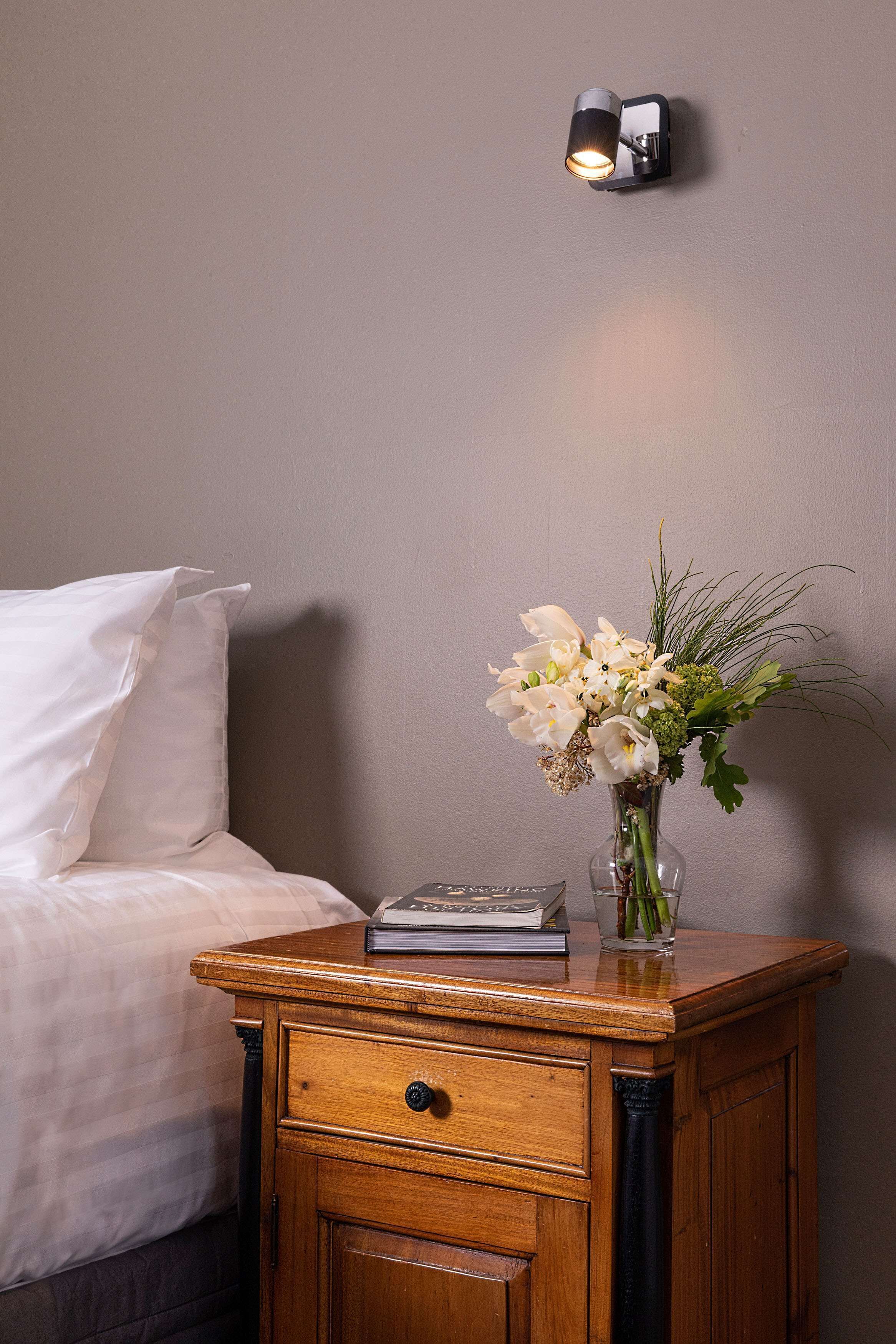 Un lit bien fait, avec du linge de lit blanc, une table de chevet ornée d’un vase de fleurs et un luminaire fixé au mur.