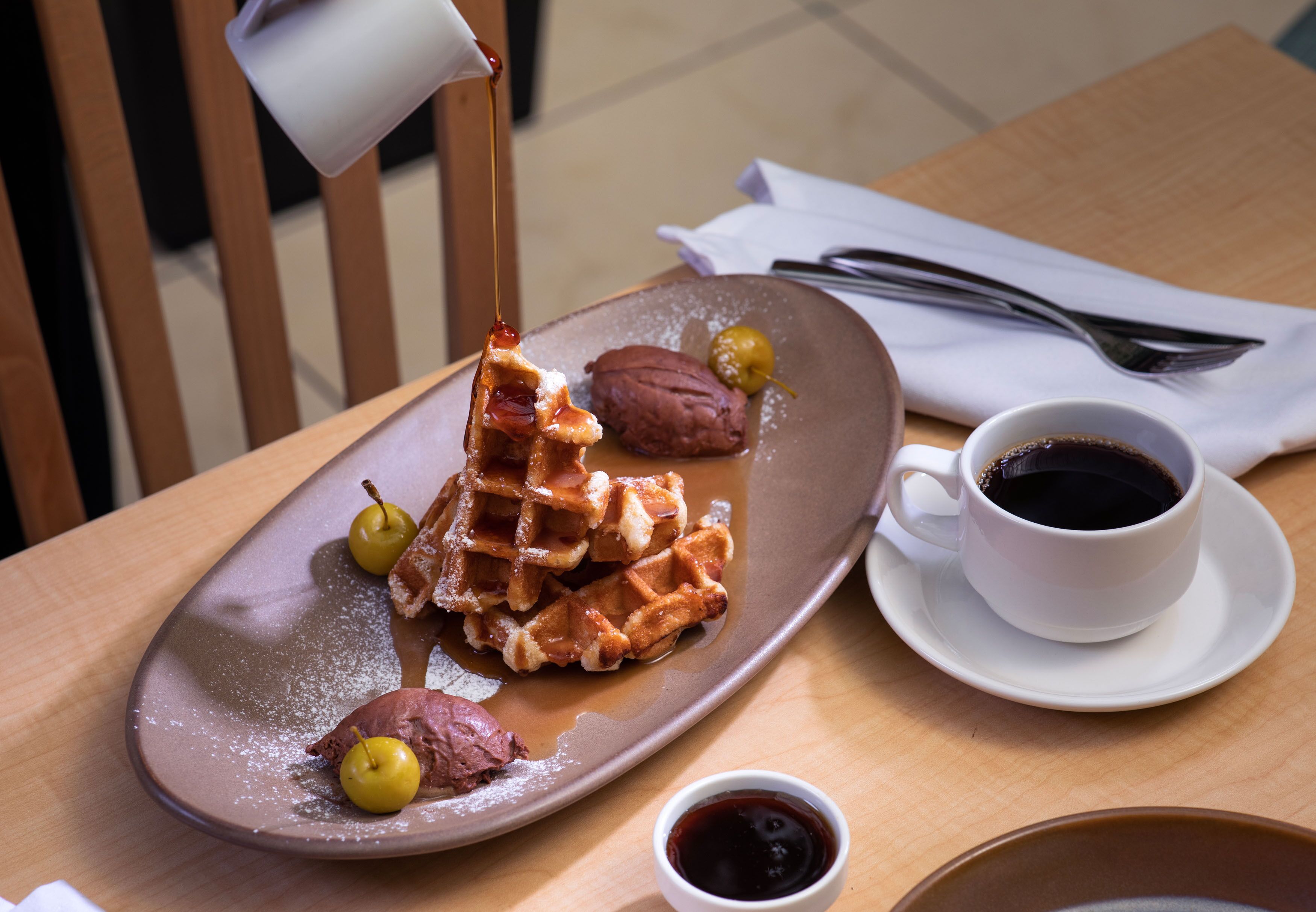 Un plato con waffles, jarabe, azúcar glas, una taza de café y un tazón pequeño de jarabe sobre una mesa de madera.
