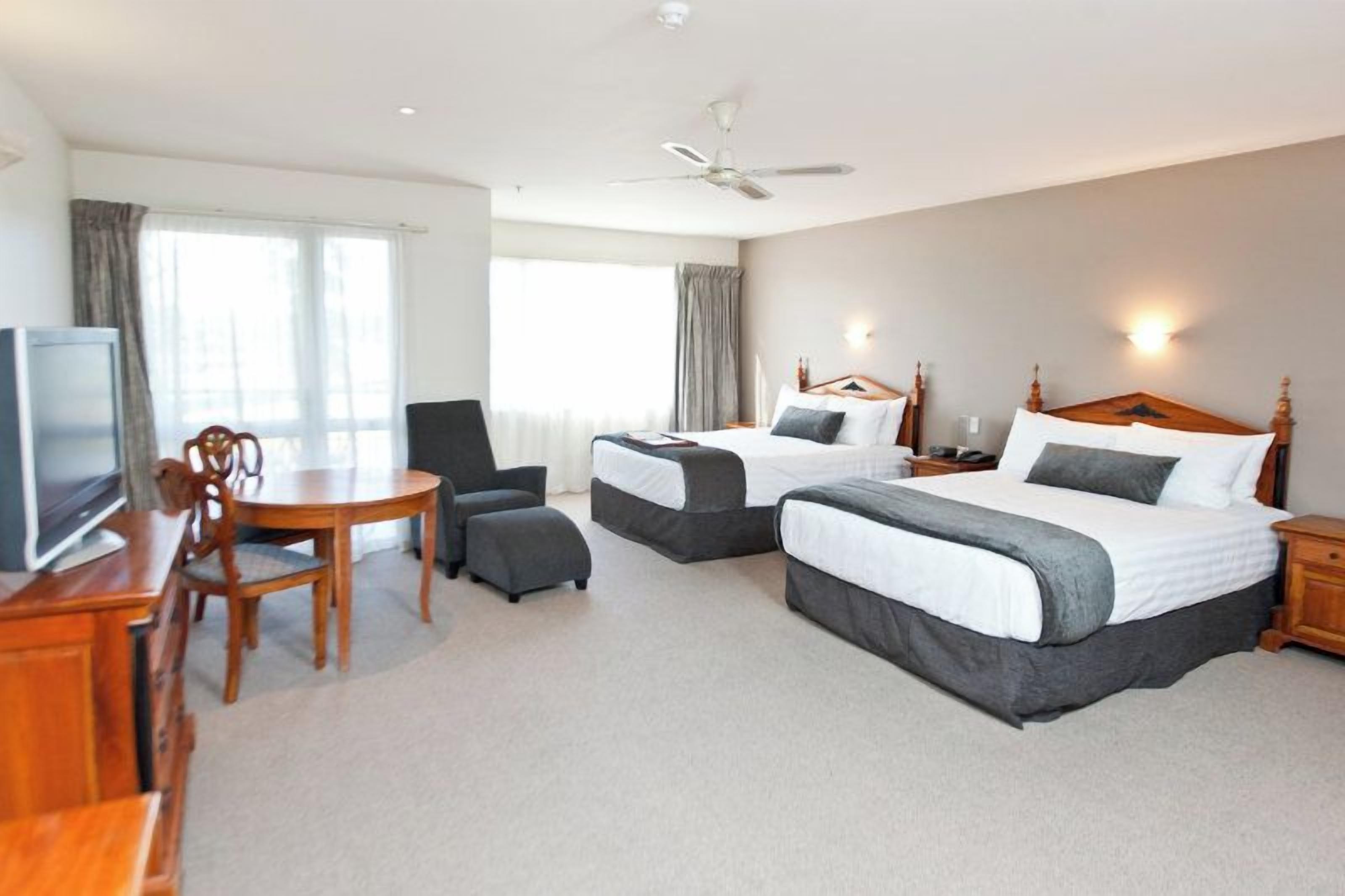 A hotel room with two beds, a desk with a chair, a television, and a ceiling fan.