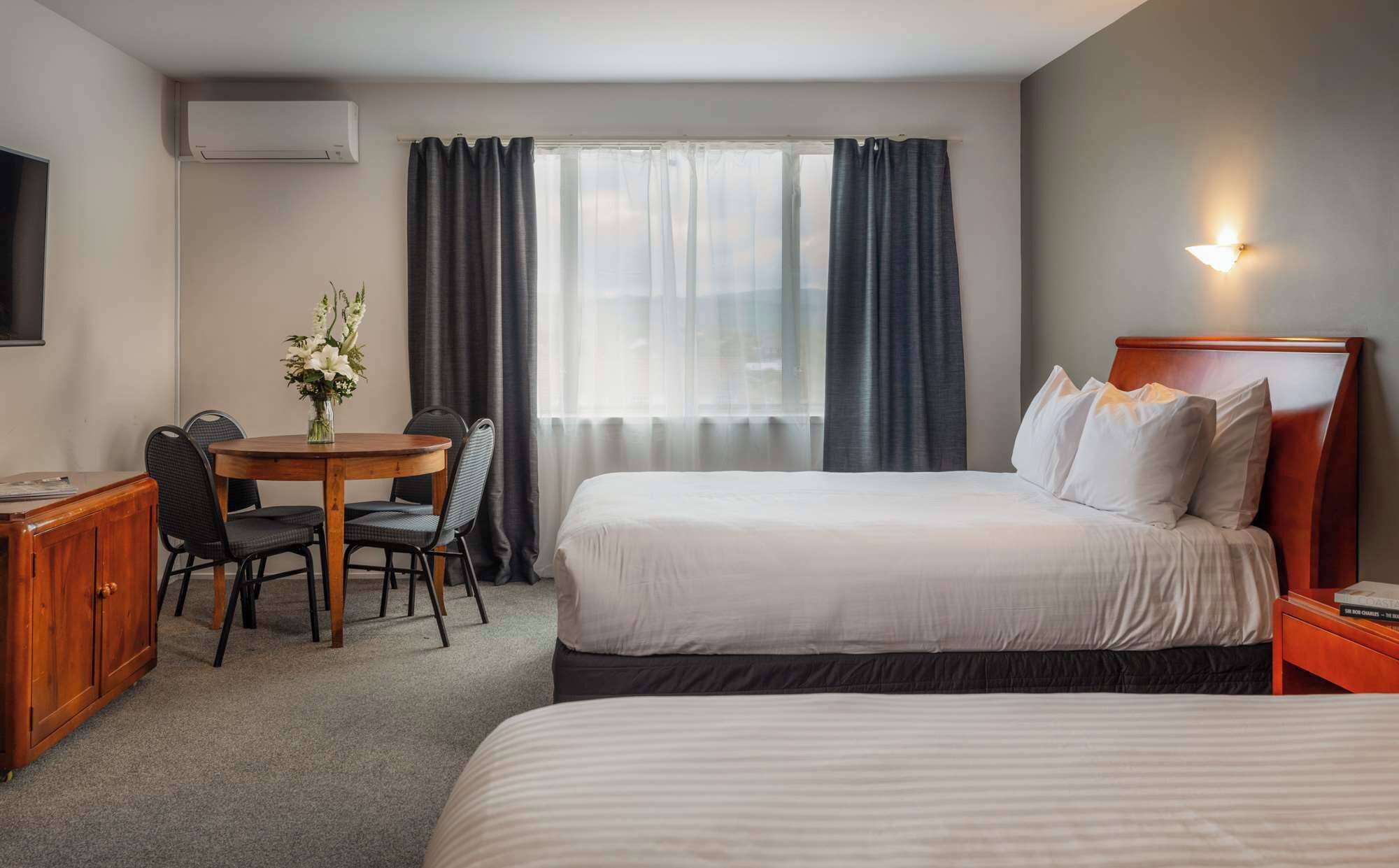 Quarto de hotel com cama, escrivaninha com cadeira, mesa pequena com um vaso de flores e televisão na parede.