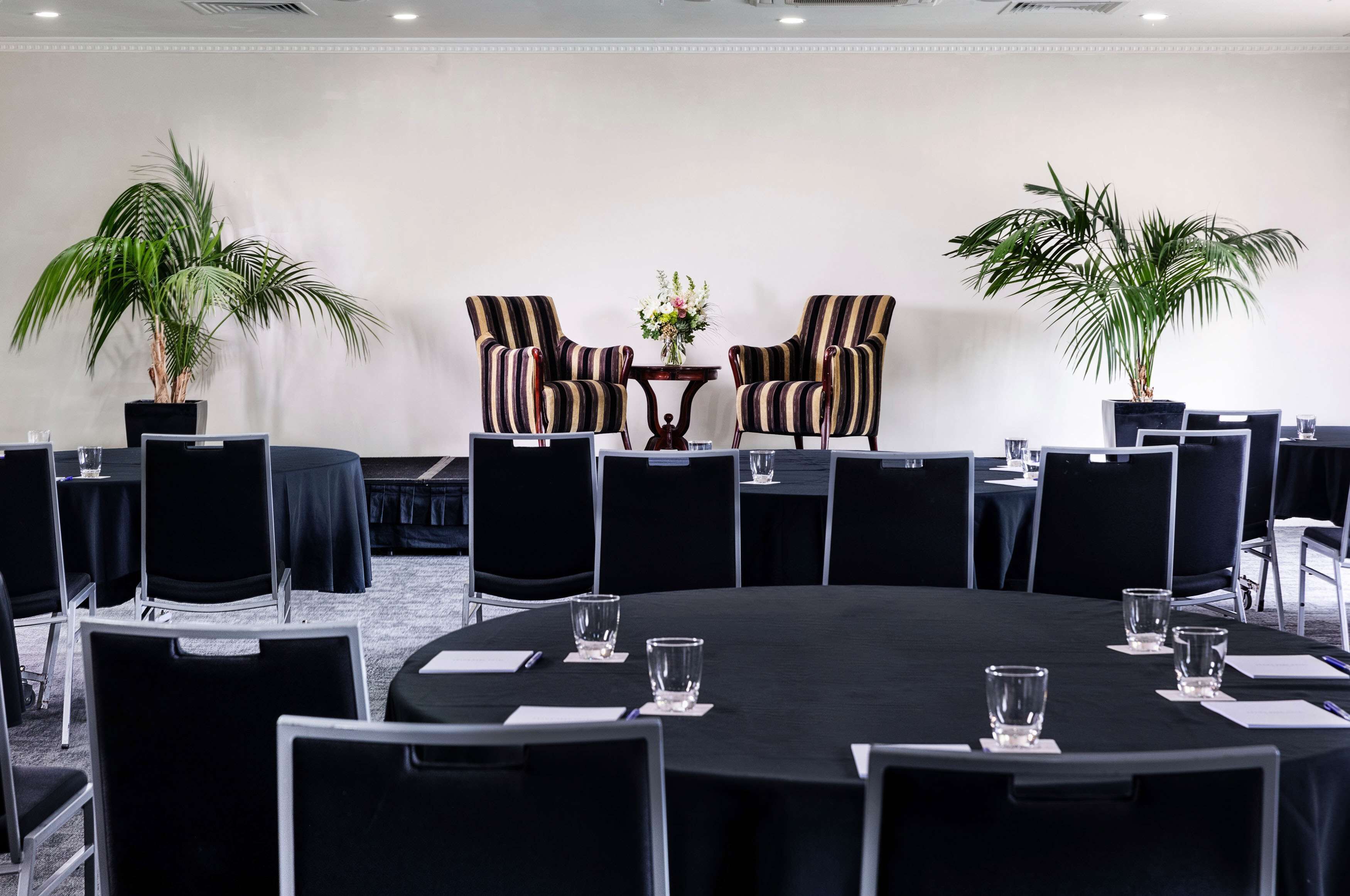Uma sala de conferências com uma mesa redonda, cadeiras pretas e duas plantas em vasos.