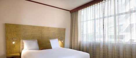 A hotel room with a large bed, two bedside lamps, a headboard, and a window with sheer curtains.