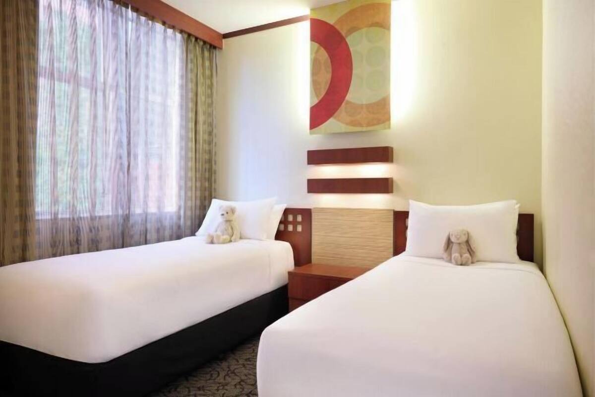 Camera d'albergo con due letti, un'ampia finestra con tende leggere, un lampadario a parete e un elemento decorativo.