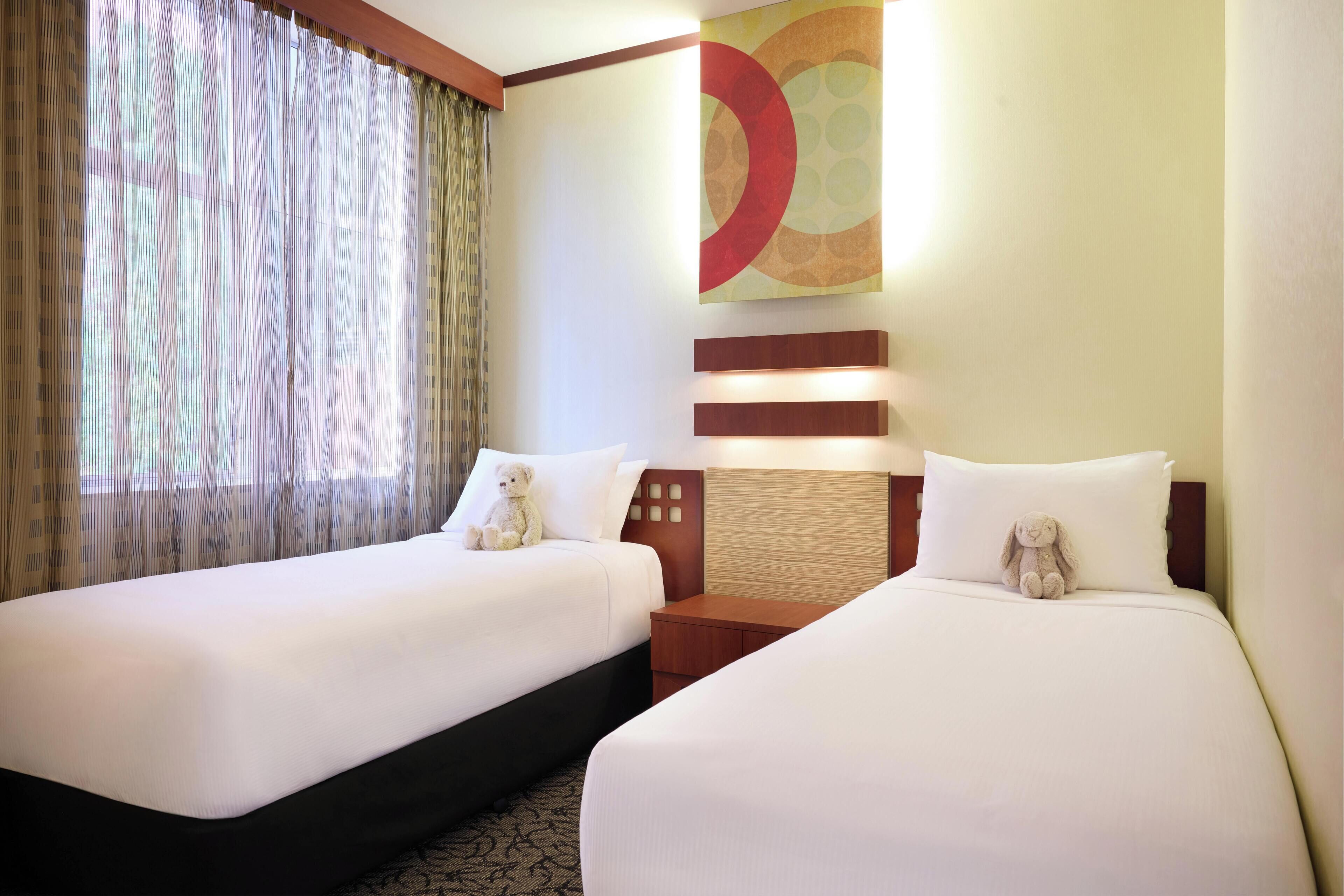 A hotel room with two beds, a large window with curtains, a wall art piece, and a nightstand with a lamp.