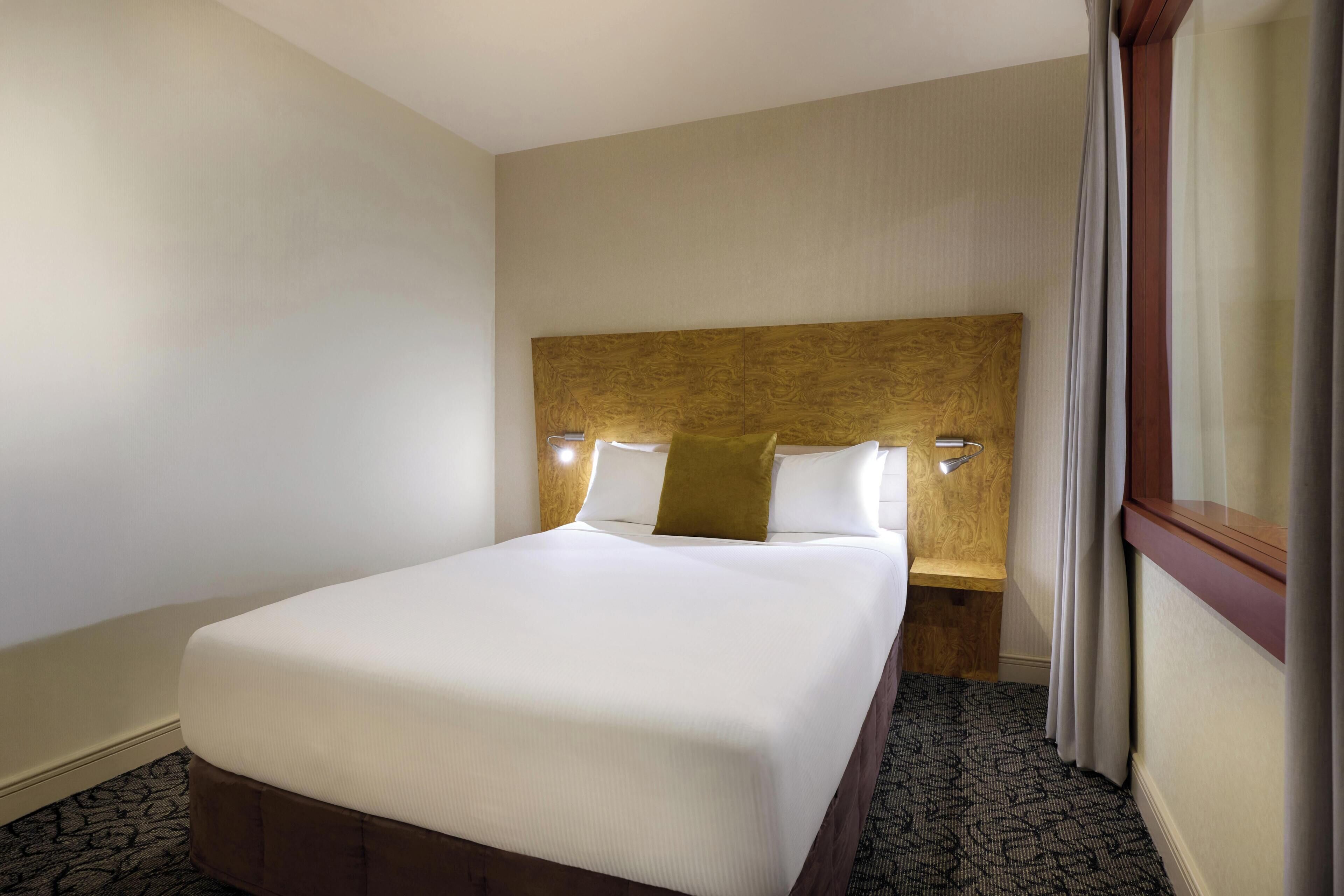 A hotel room with a bed, a headboard with wall-mounted lights, a wooden nightstand, and a patterned carpet.