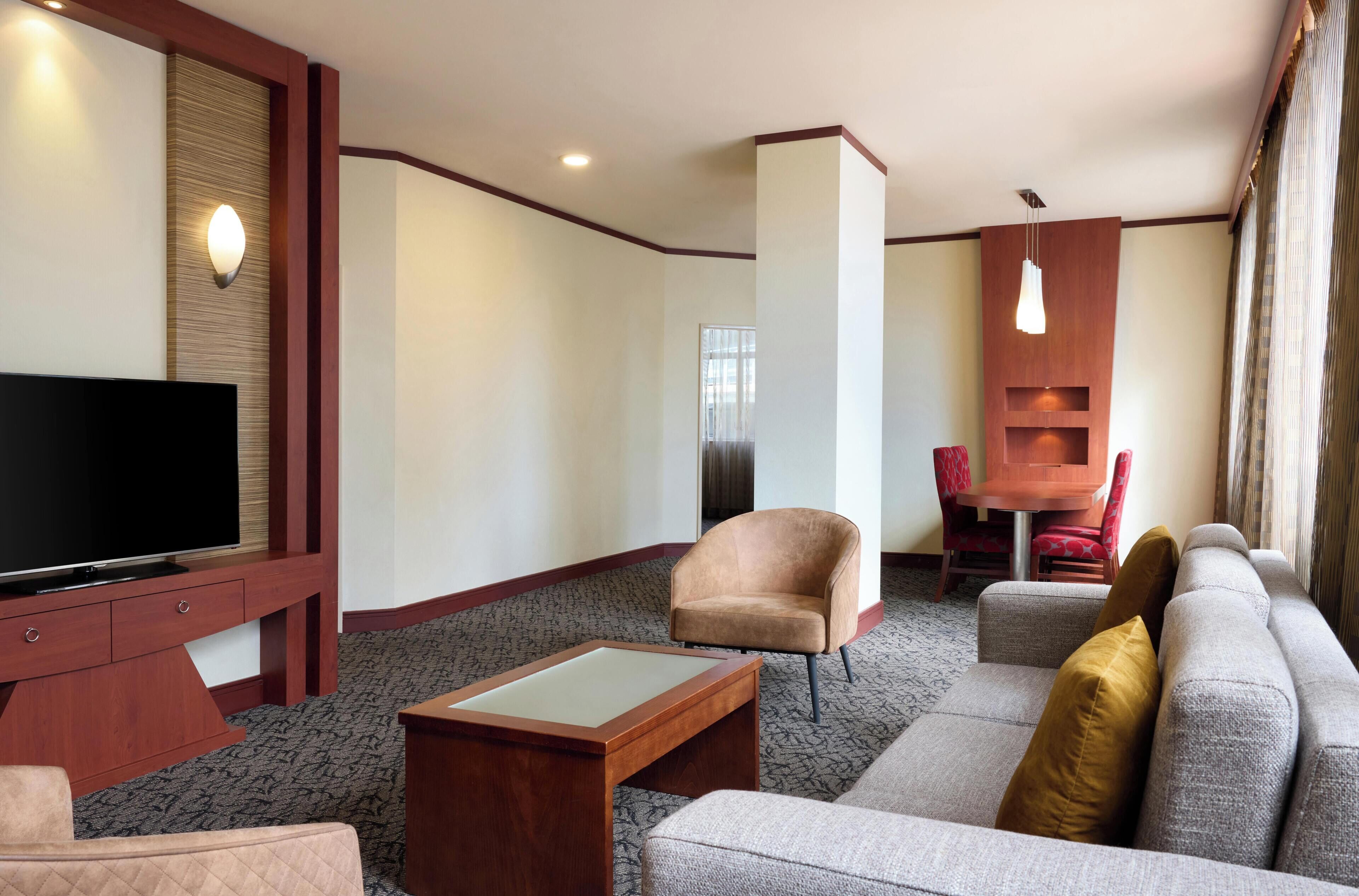 A hotel room with a flat-screen TV, a sofa, a chair, a small table, and a dining area with a table and chairs.