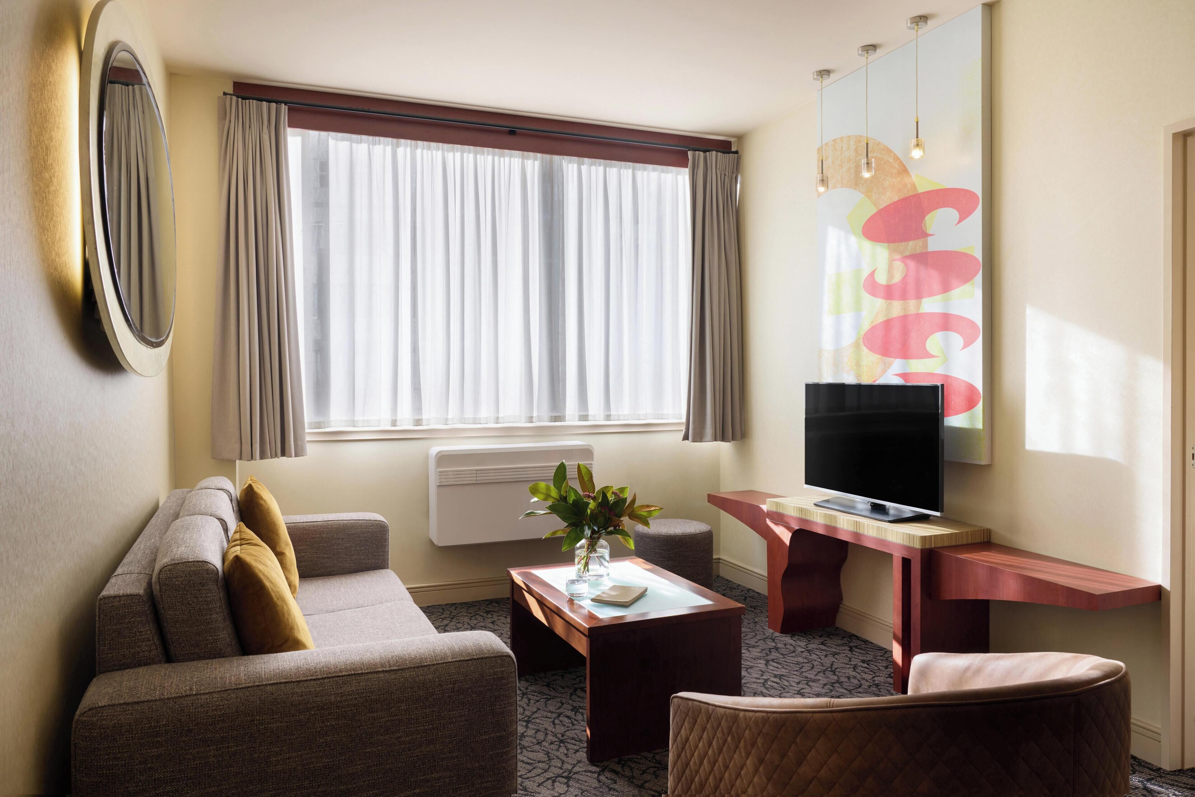 A hotel room with a sofa, a small table with a vase, a television on a wooden stand, and a large window with curtains.