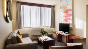 Una habitación de hotel con sofá, una mesita con un jarrón, televisor sobre un mueble de madera y un amplio ventanal con cortinas.