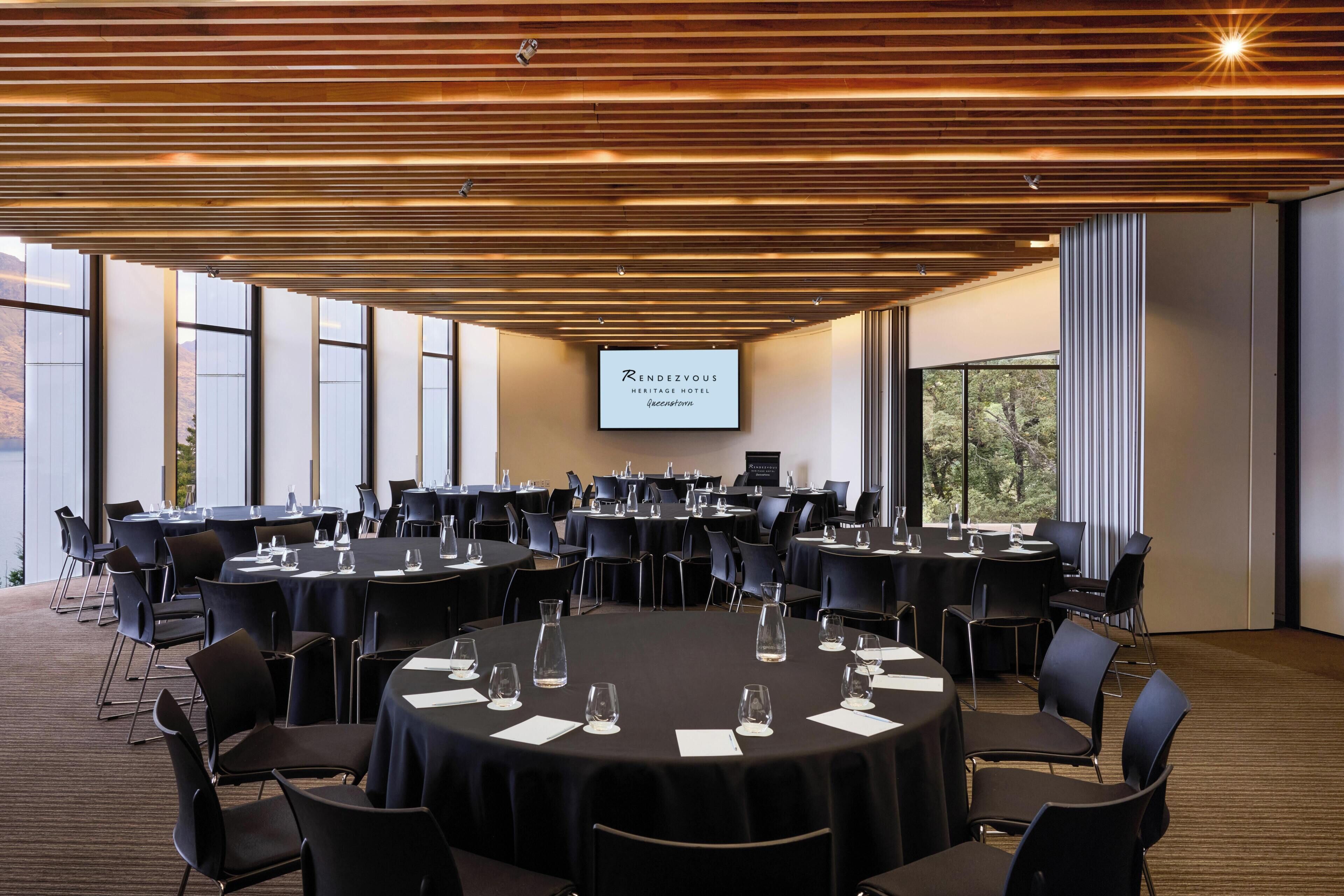 A conference room with round tables, black chairs, and a large screen displaying the hotel name.