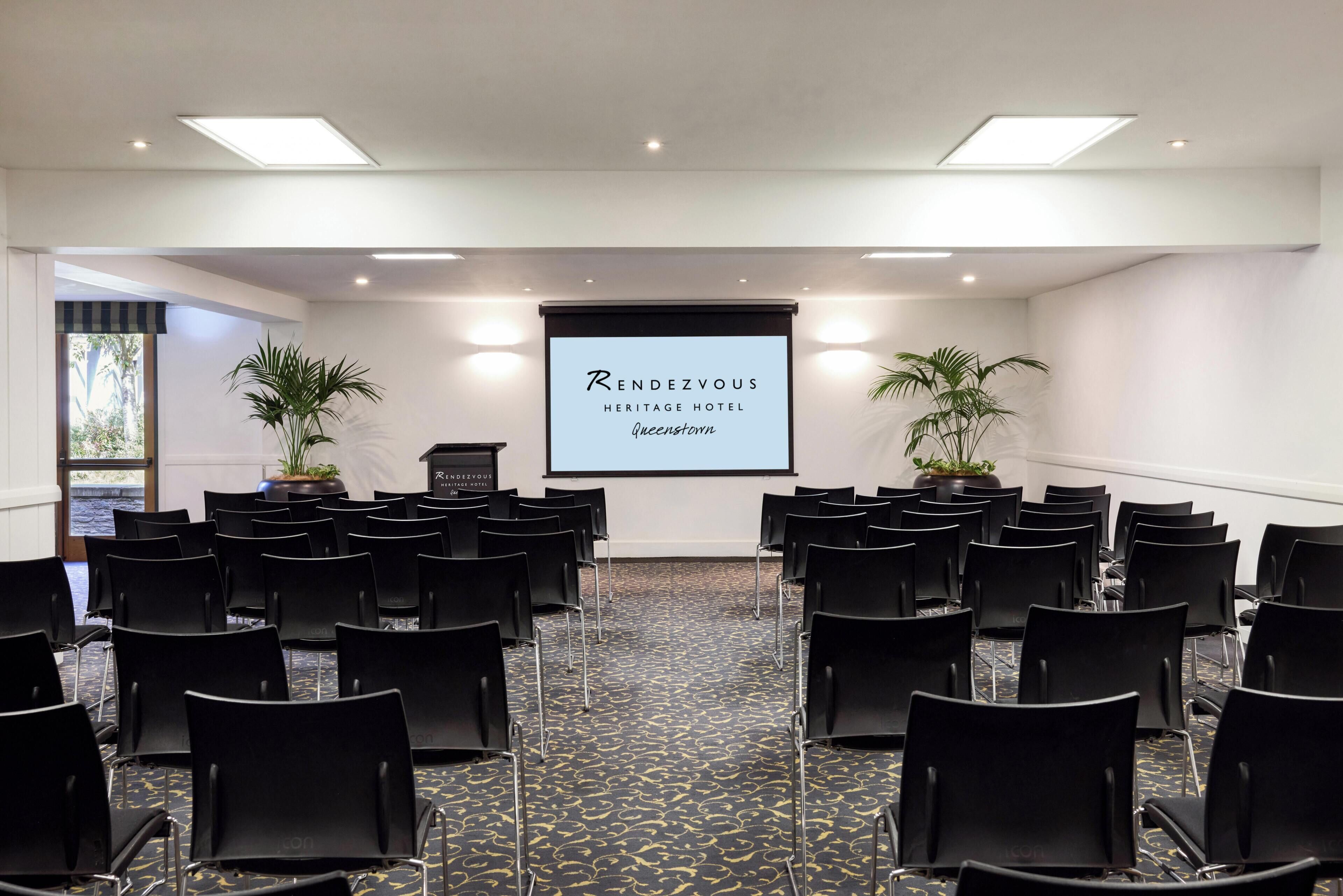 Un salón de conferencias con filas de sillas negras, una pantalla de proyector mostrando 'Rendezvous Heritage Hotel Queenstown' y dos plantas en macetas.