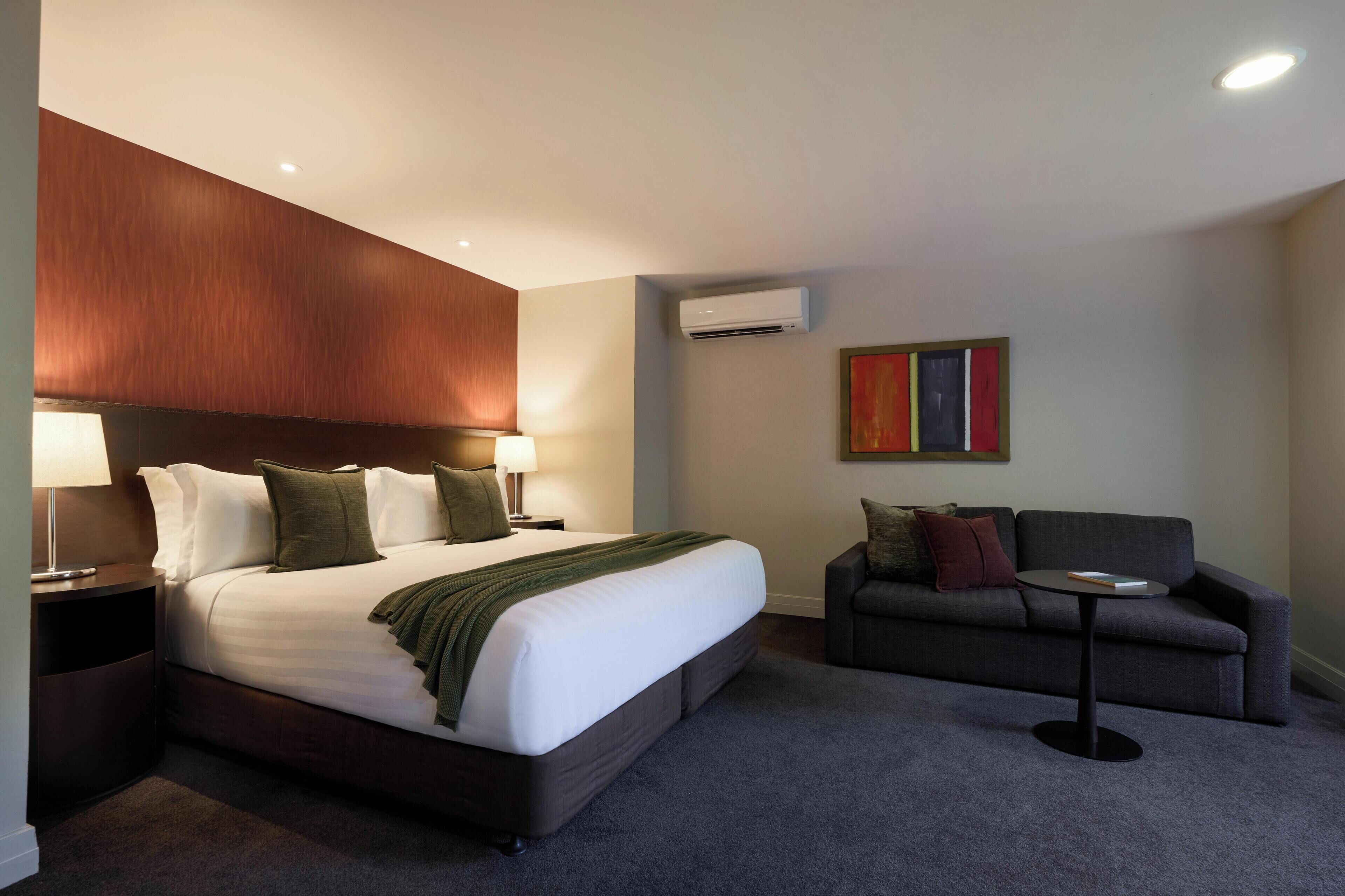 A hotel room with a bed, a sofa, a small table, an air conditioner, and a painting on the wall.