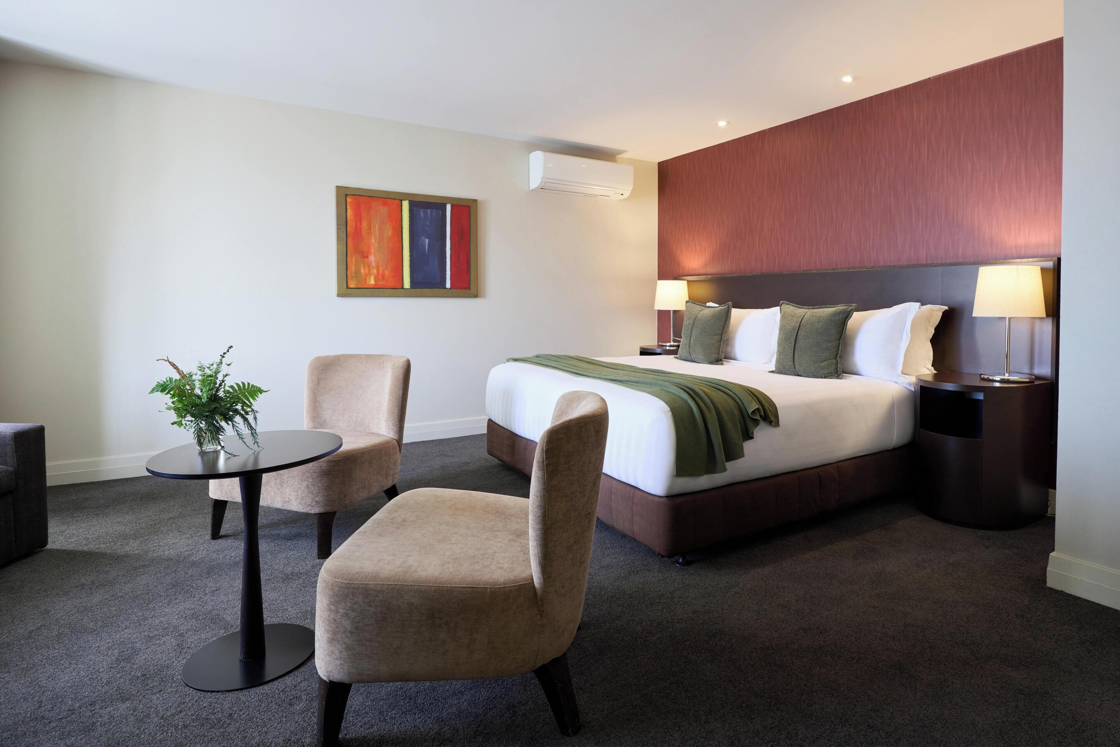 Una camera d'albergo con un letto, due poltrone, un tavolino, una pianta, un quadro e un comodino con una lampada.