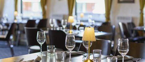 A dining area with tables set for a meal, featuring a lamp, wine glasses, and cutlery.
