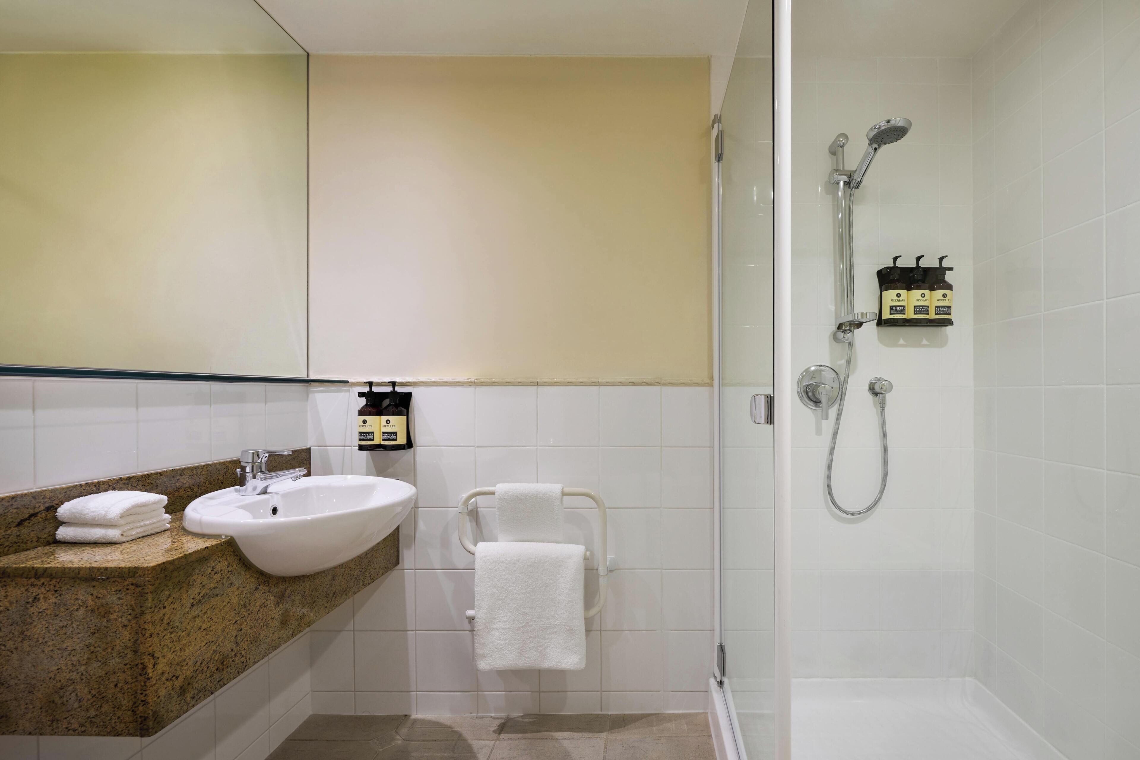 A bathroom with a sink, mirror, towel rack, shower, and toiletries.