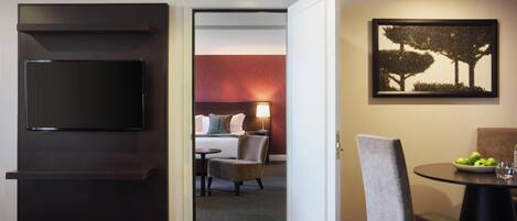 A hotel room with a flat-screen TV, a dining area with a table and chairs, and a painting on the wall.