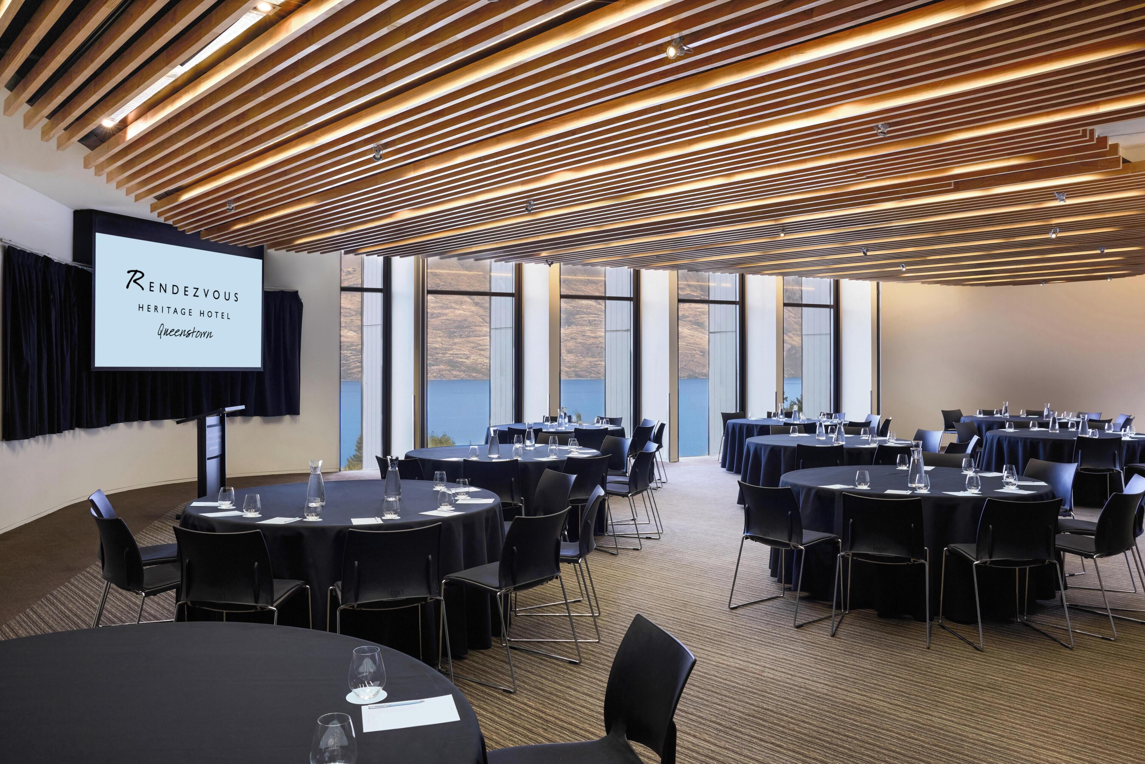 Un salón de conferencias con varias mesas y sillas, una pantalla de proyector mostrando 'Rendezvous Heritage Hotel Queenstown' y amplias ventanas que ofrecen vistas al exterior.