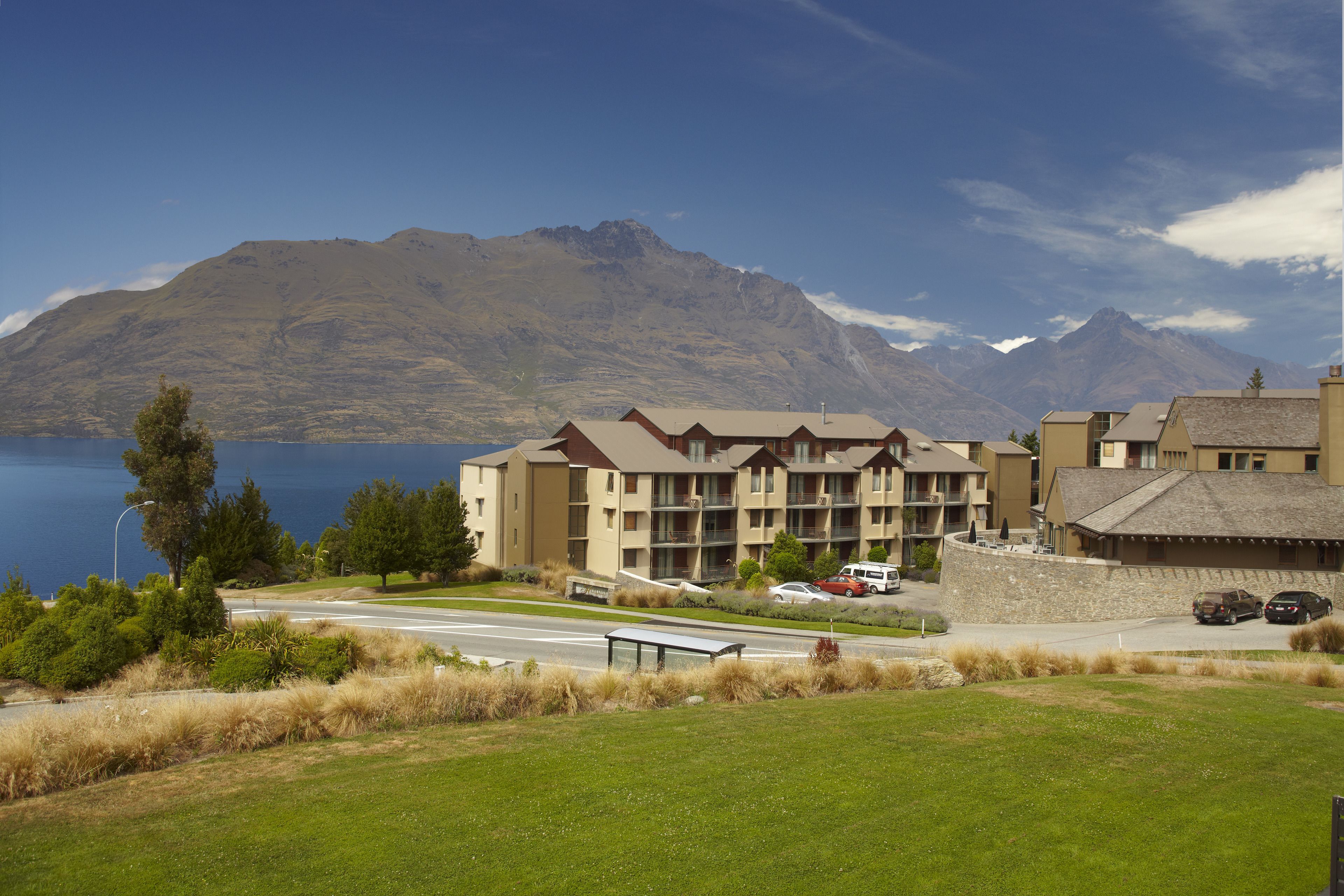 A hotel with multiple floors, a parking area, and a view of mountains and a lake.