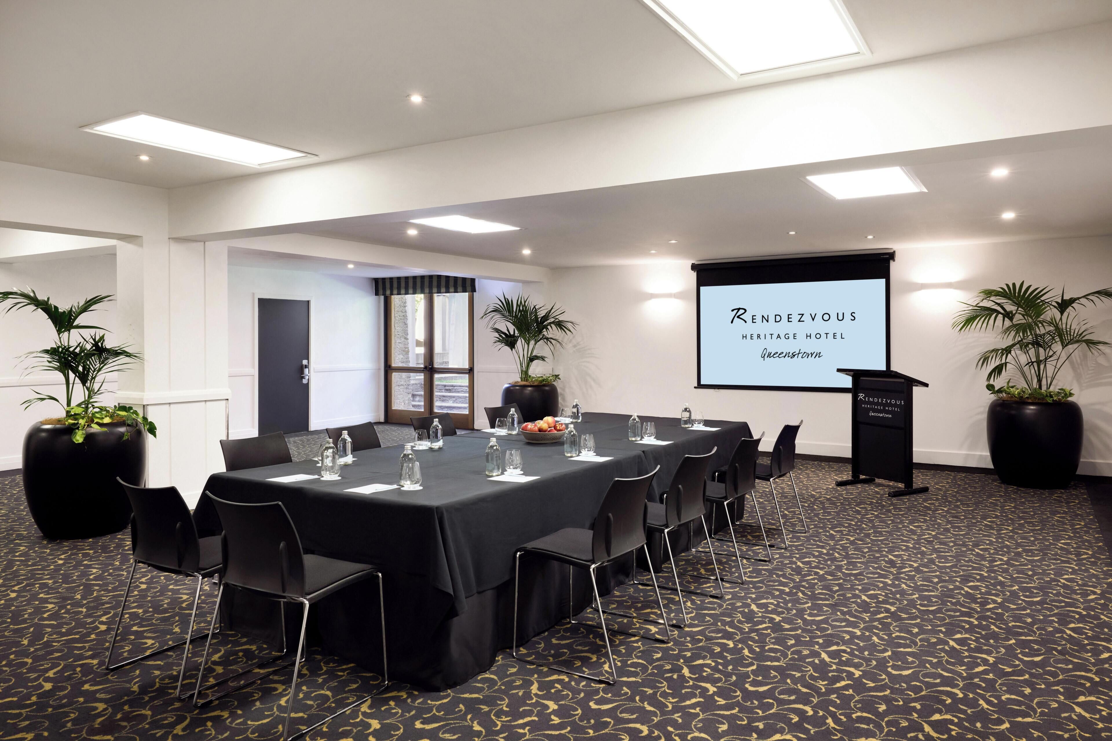 A conference room with a long table, chairs, a projector screen, and potted plants.