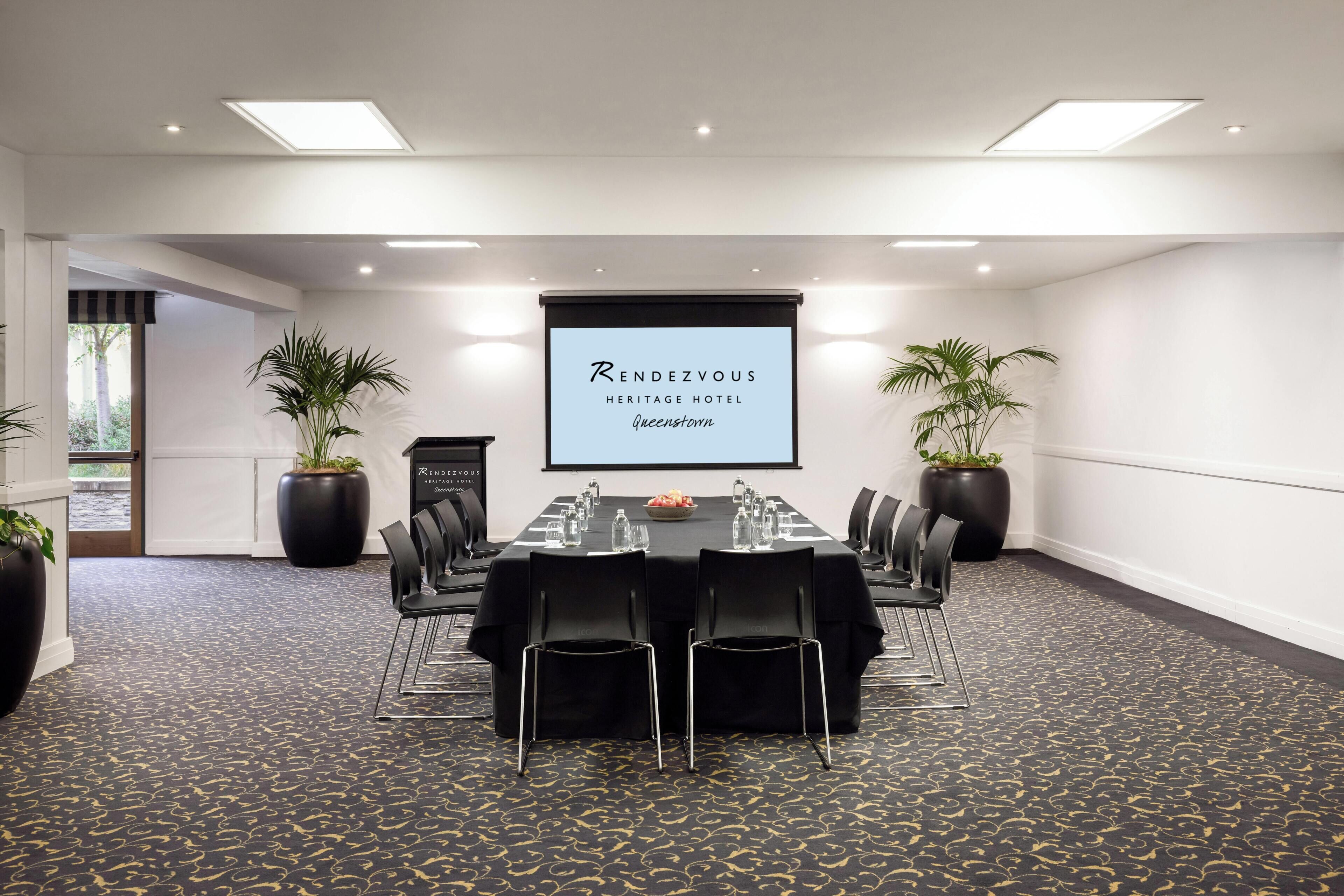 A conference room at the Rendezvous Heritage Hotel in Queensland with a projector screen, a long table, chairs, and potted plants.