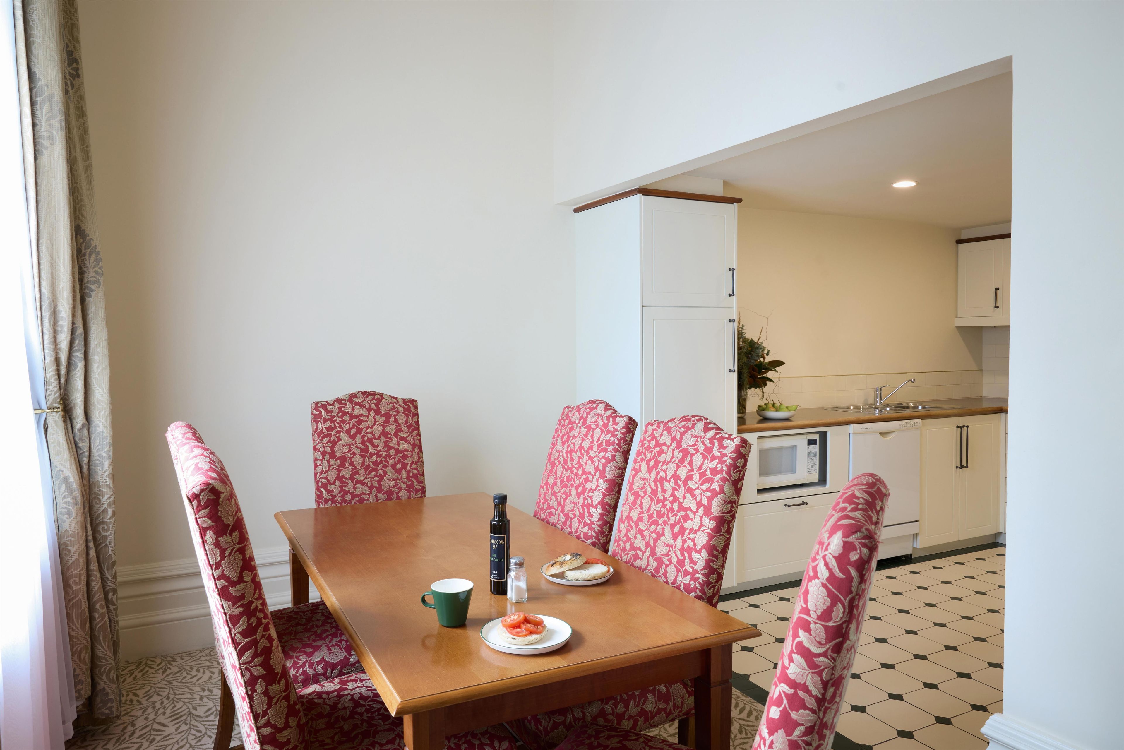 A dining area with a wooden table, floral-patterned chairs, and a view into a kitchen with a microwave and cabinets.