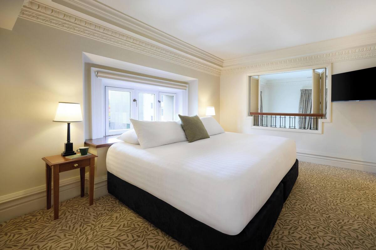 Ein Hotelzimmer mit einem großen Bett, einem Nachttisch mit Lampe, einem Fenster und einem an der Wand befestigten Fernseher.