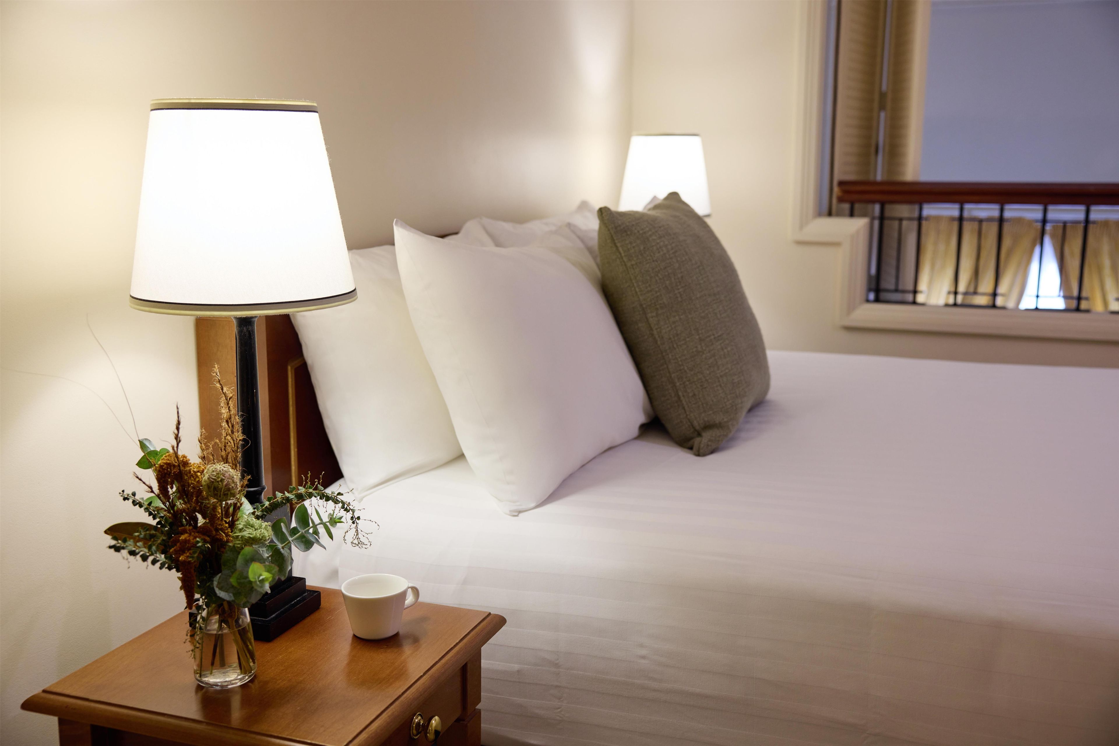 Habitación de hotel con una cama bien tendida, una mesita de noche con una lámpara, un florero con flores secas y una taza.