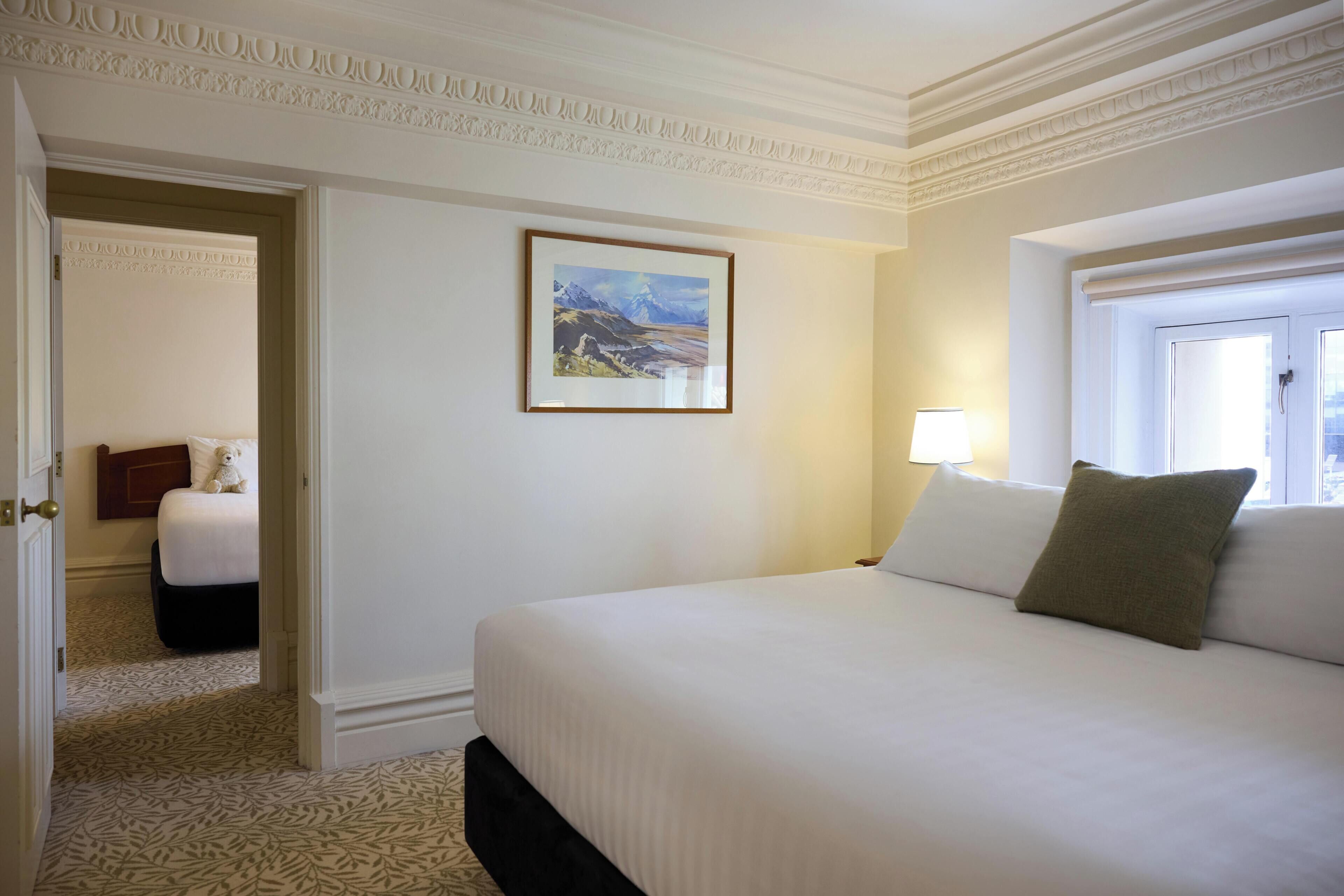 Een hotelkamer met een bed, een schilderij aan de muur, een raam en een deur die leidt naar een andere kamer.
