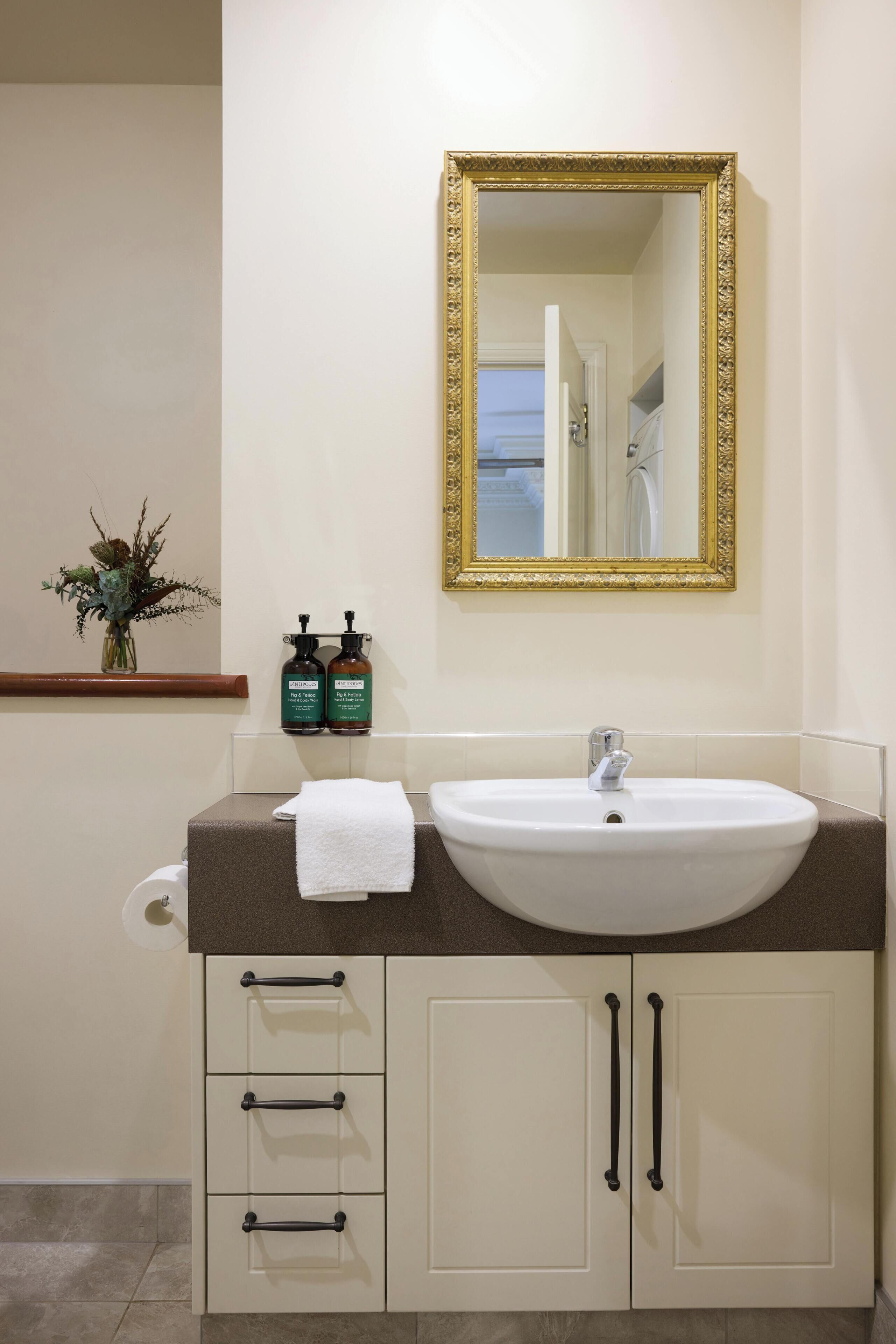 A bathroom with a white sink, a gold-framed mirror, and a vase with dried flowers on the wall.