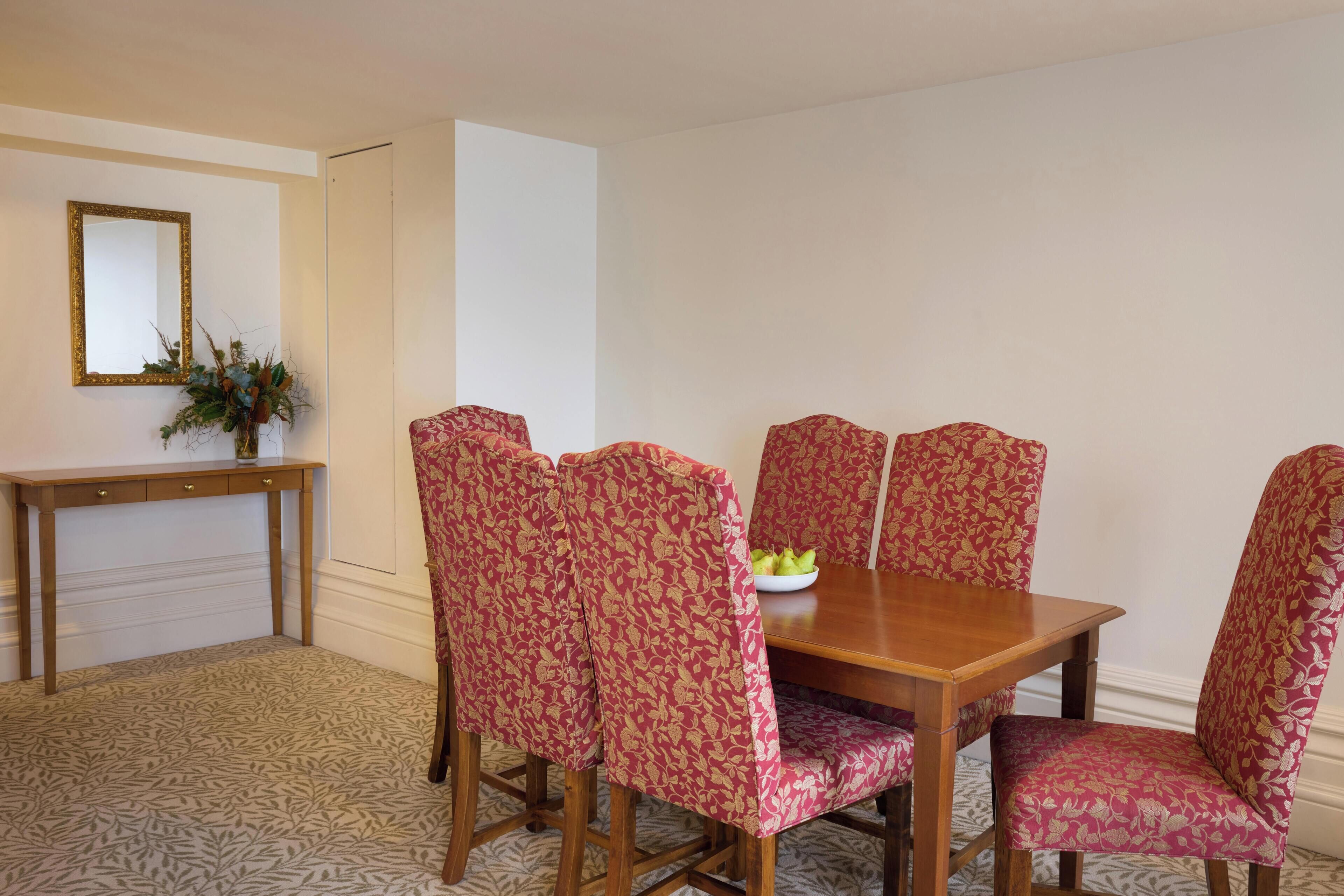 Un coin repas avec une table en bois et des chaises à motifs floraux, un miroir au mur et une console sur laquelle est posé un vase rempli de verdure.