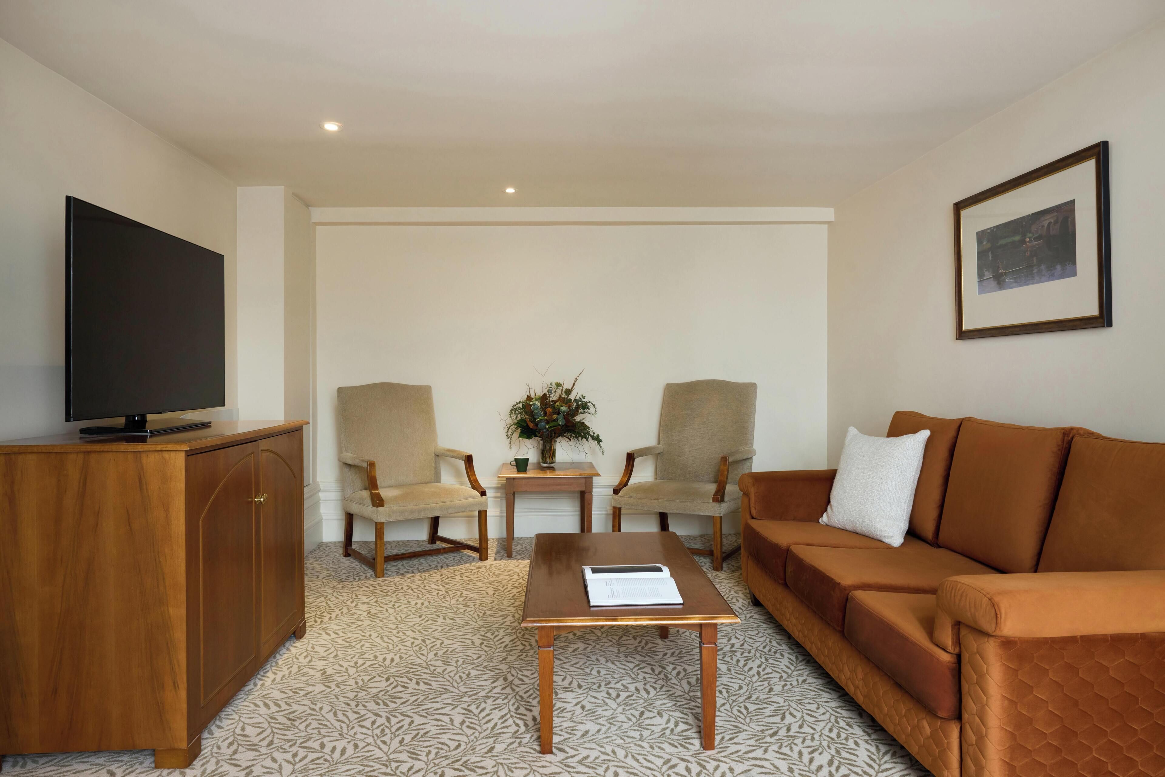 Un salon comprenant un canapé, deux fauteuils, une table basse, un téléviseur posé sur un meuble en bois, un tableau encadré accroché au mur, et une petite table sur laquelle se trouvent un vase et une plante.