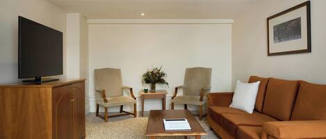Un salon comprenant un canapé, deux fauteuils, une table basse, un téléviseur posé sur un meuble en bois, un tableau encadré accroché au mur, et une petite table sur laquelle se trouvent un vase et une plante.