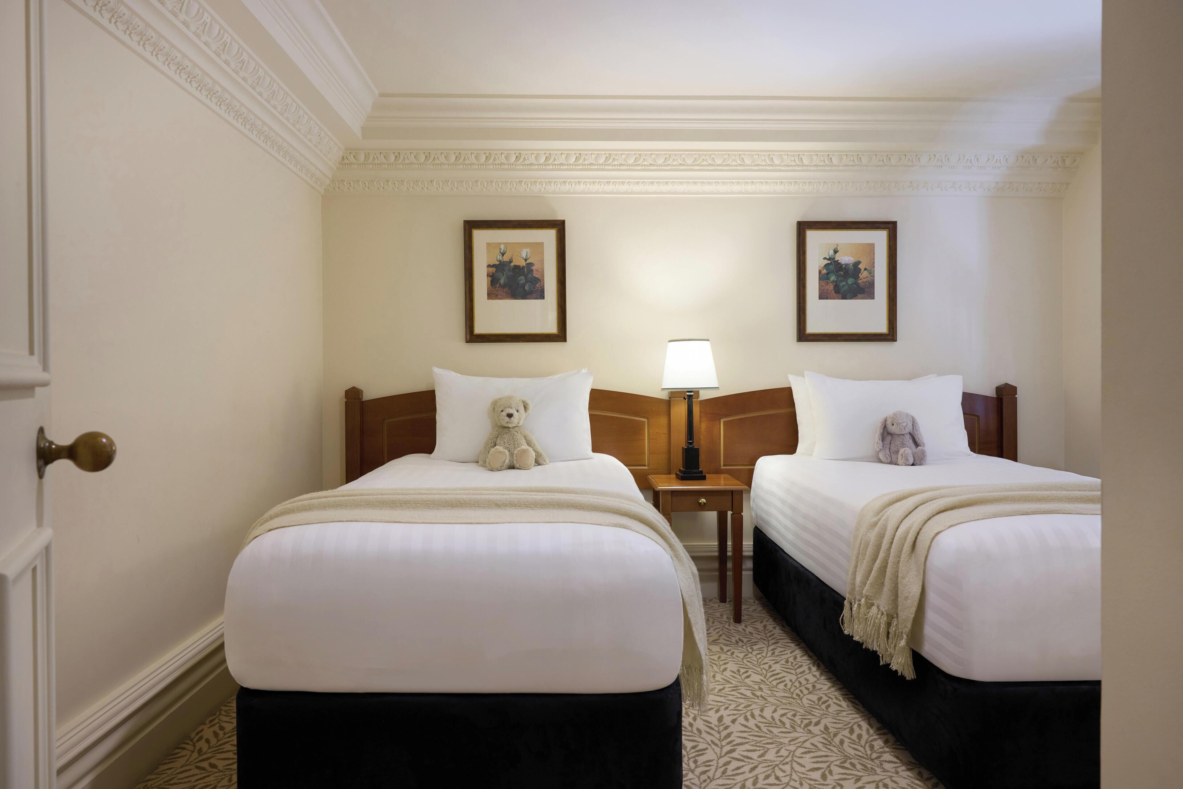 Een hotelkamer met twee bedden, een deur, een nachtkastje met een lamp en twee ingelijste foto's aan de muur.