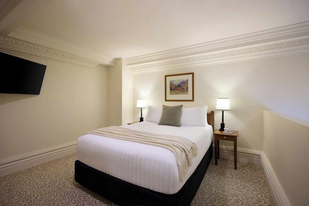 Habitación de hotel con una cama grande, dos mesitas de noche con lámparas, un televisor de pantalla plana en la pared y un cuadro enmarcado sobre la cama.
