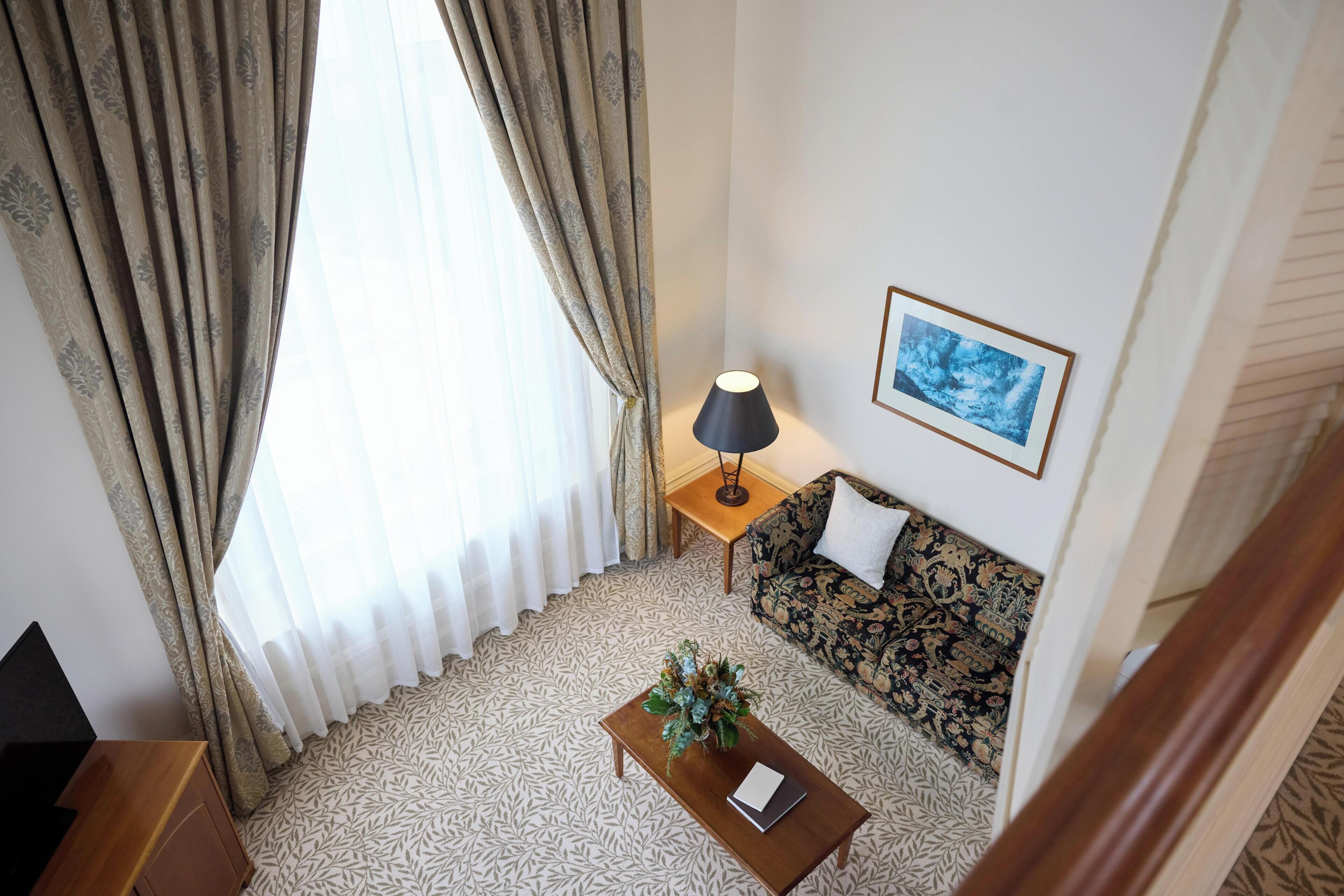 A room with a patterned floor, a sofa with a floral design, a small table with a lamp, and a framed picture on the wall.