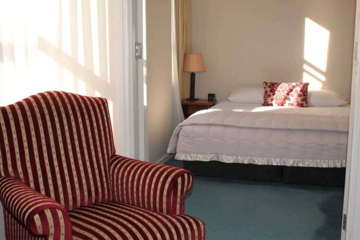 Une chambre d’hôtel avec un fauteuil à rayures, un lit avec une literie blanche, une table de chevet avec une lampe, et une vue sur l’extérieur par la fenêtre.