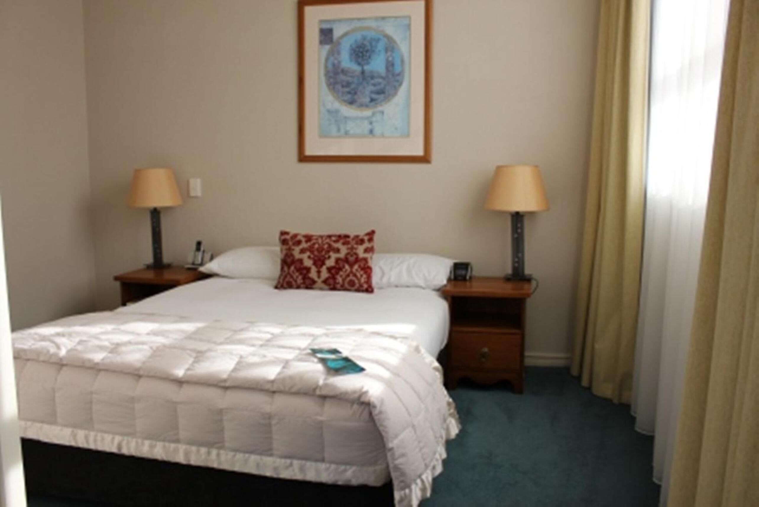 A neatly made bed with a quilt, two bedside lamps, a framed artwork on the wall, and a window with curtains.