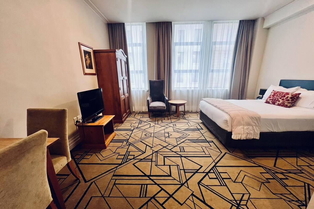 Une chambre d’hôtel avec un lit, un téléviseur posé sur un meuble en bois, une chaise, une petite table et un tapis à motifs.