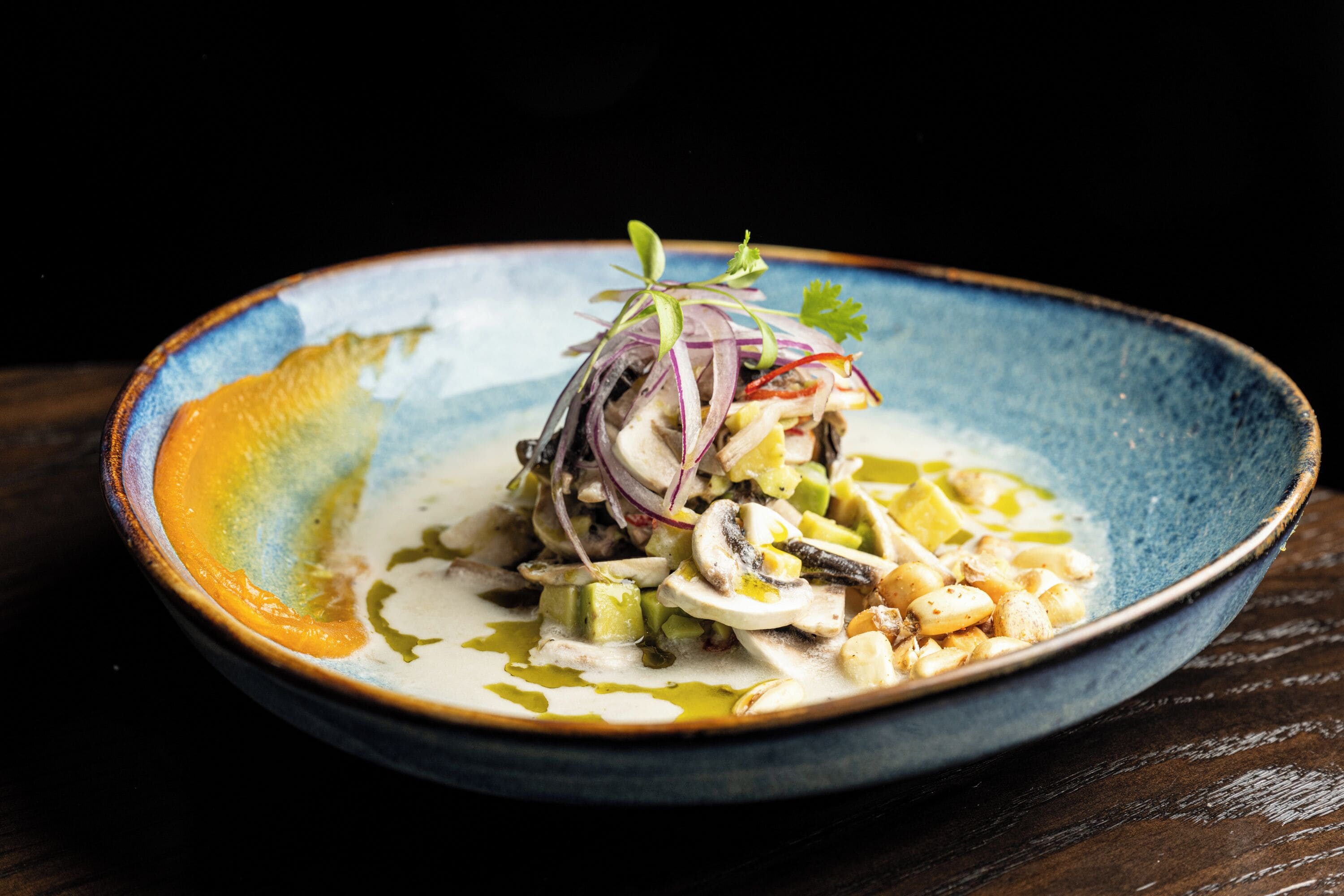 A blue ceramic plate with a seafood dish, garnished with red onion and herbs, on a dark wooden table.