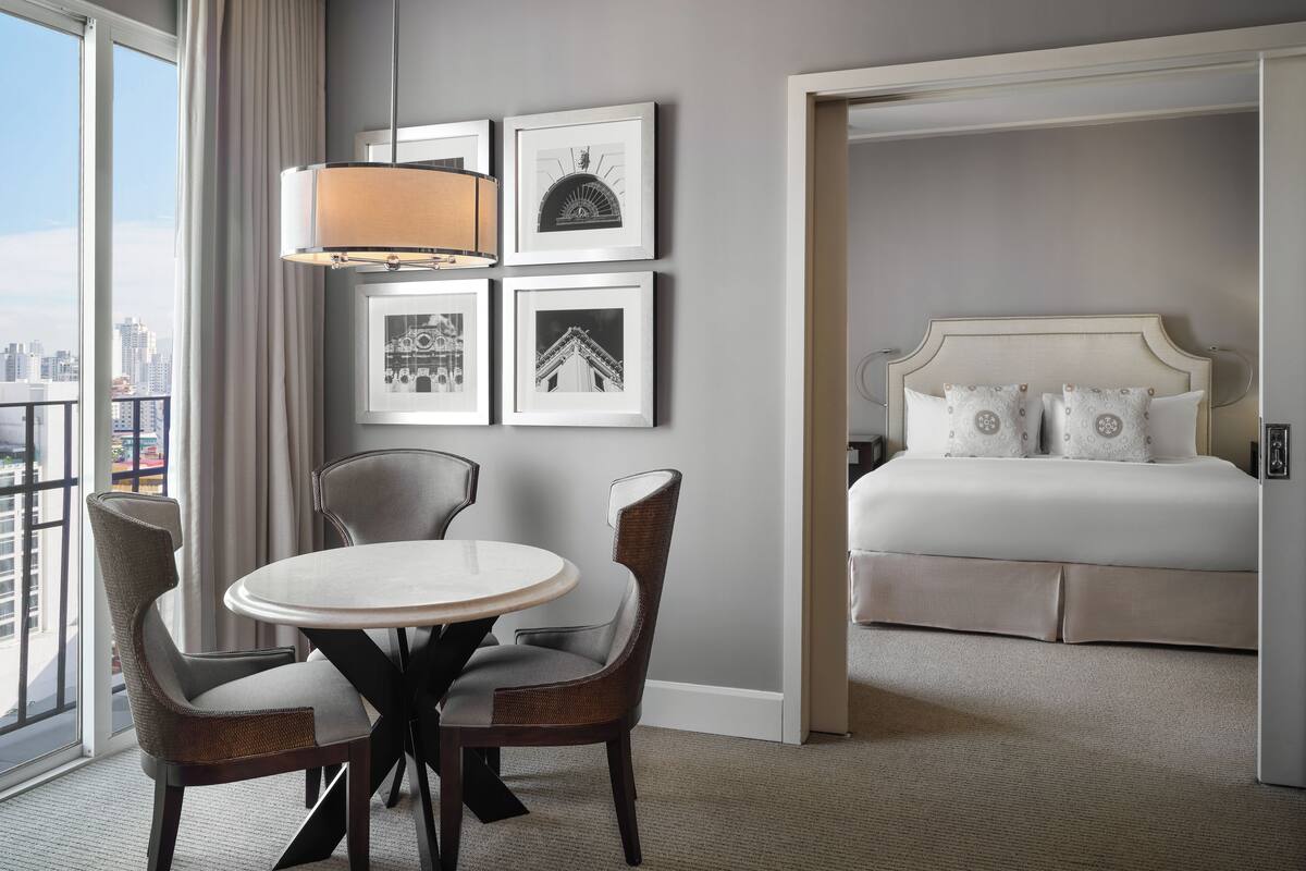 Een hotelkamer met een ronde eettafel, twee stoelen, een bed met hoofdeinde, en ingelijste foto's aan de muur.