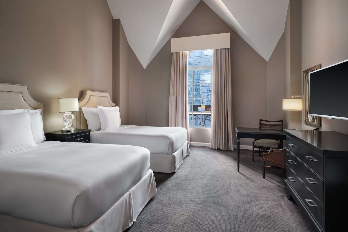 Een hotelkamer met twee bedden, een bureau, een dressoir en een groot raam met gordijnen.