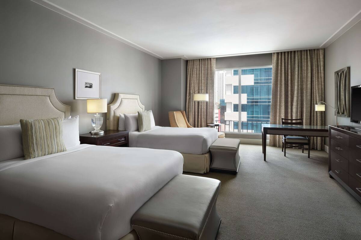 Een hotelkamer met twee bedden, een bureau, een stoel en een groot raam met gordijnen.