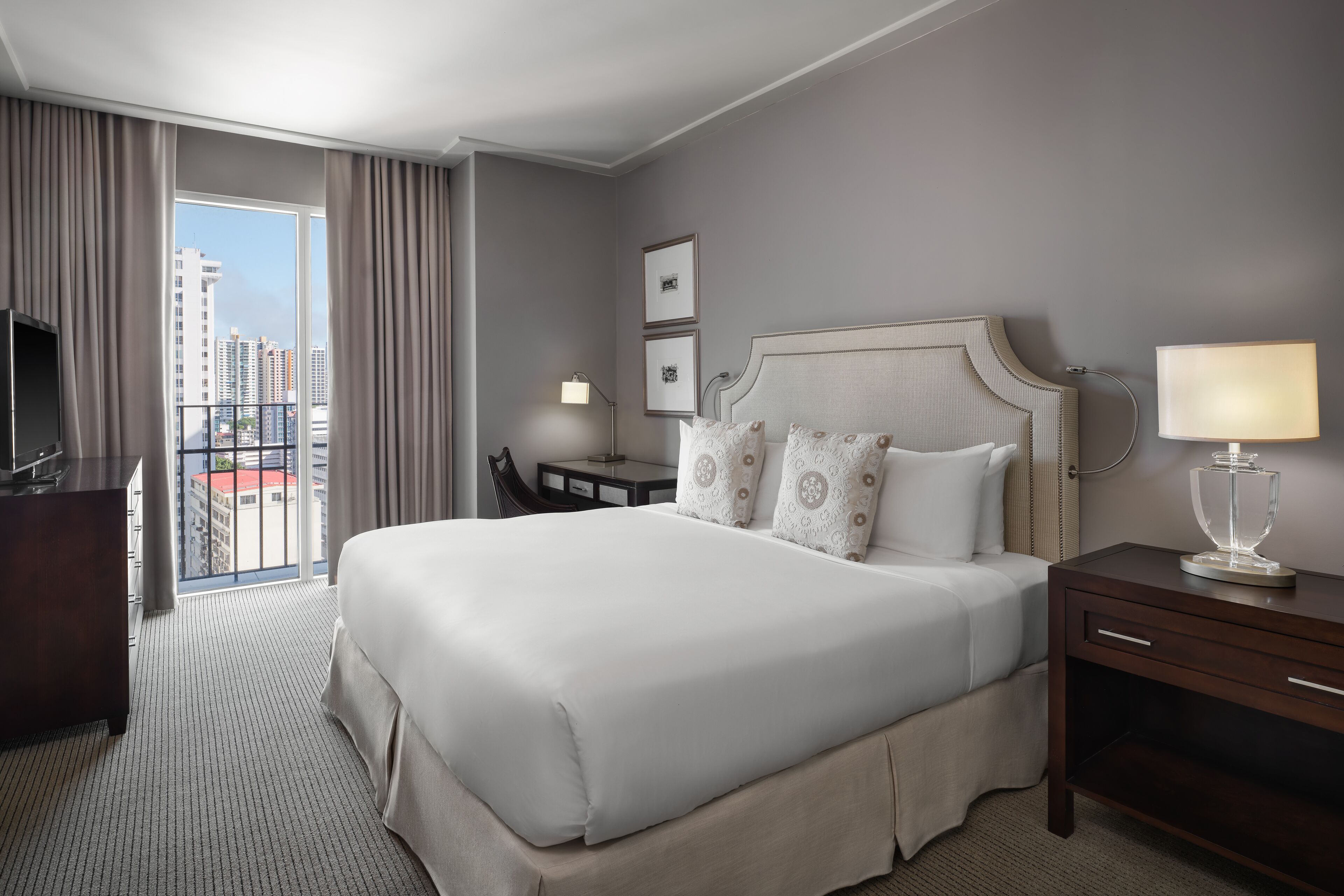 A hotel room with a large bed, a desk, a chair, a TV, and a view of the city through the window.