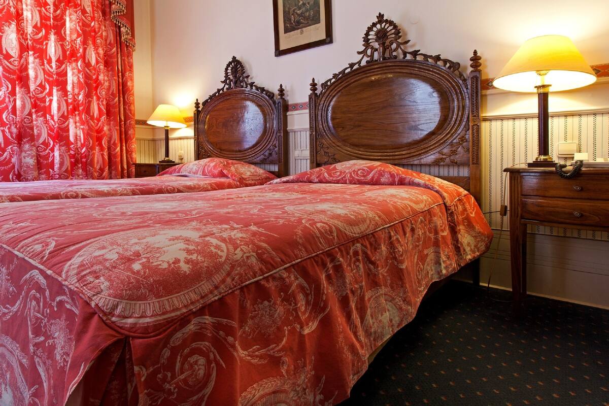A hotel room with two beds, red patterned bedspreads, wooden headboards, a nightstand with a lamp, and a wall-mounted telephone.