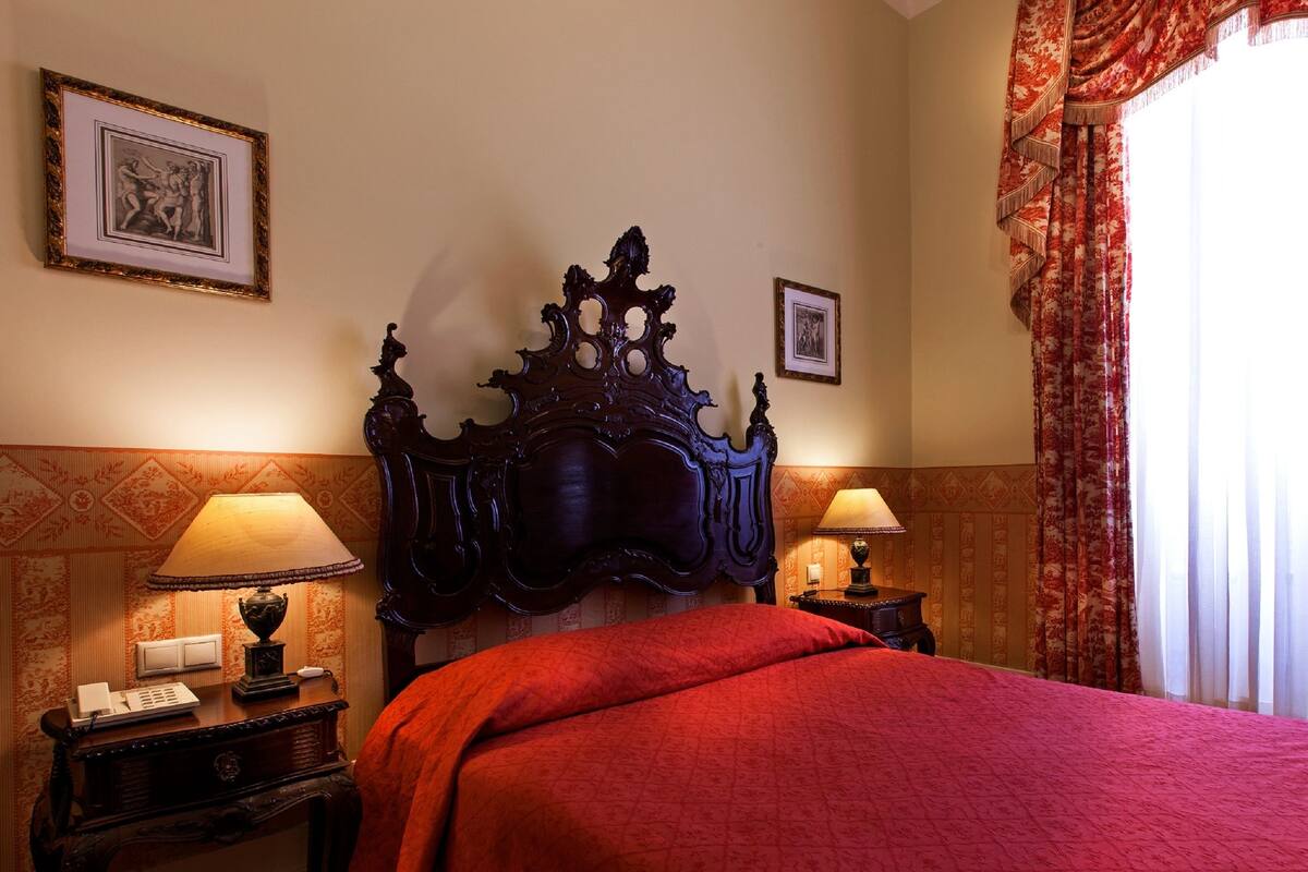 A hotel room with a large ornate bed, two bedside tables with lamps, a framed picture on the wall, and a window with curtains.